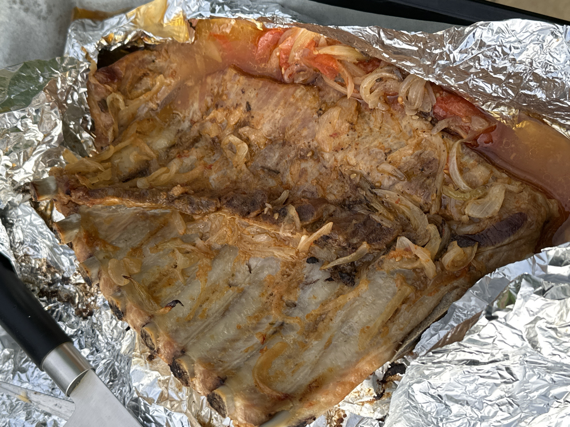 This close-up, high-angle shot captures a large portion of cooked meat, likely pork ribs, served within a crumpled aluminum foil container. The meat, showing clear bone structure, is golden-brown with some areas appearing slightly charred, indicating thorough cooking. Scattered over the meat are generous amounts of softened, translucent sliced onions and pieces of cooked tomato, which have melded into a glistening, savory liquid accumulated at the bottom of the foil. A knife with a black handle and a silver blade is partially visible in the bottom-left corner, suggesting the dish is ready for carving or serving. The aluminum foil, stained with cooking juices and fats, serves as a rustic baking or serving vessel. A dark, possibly black, edge of a baking tray or surface is visible along the top edge of the frame. The image focuses entirely on the food, conveying a sense of a recently prepared meal. There are no people visible, nor are there any explicit cues for time of day, weather, or emotions. No visible text is present in the image. The scene depicts a prepared meal, potentially from a domestic setting.