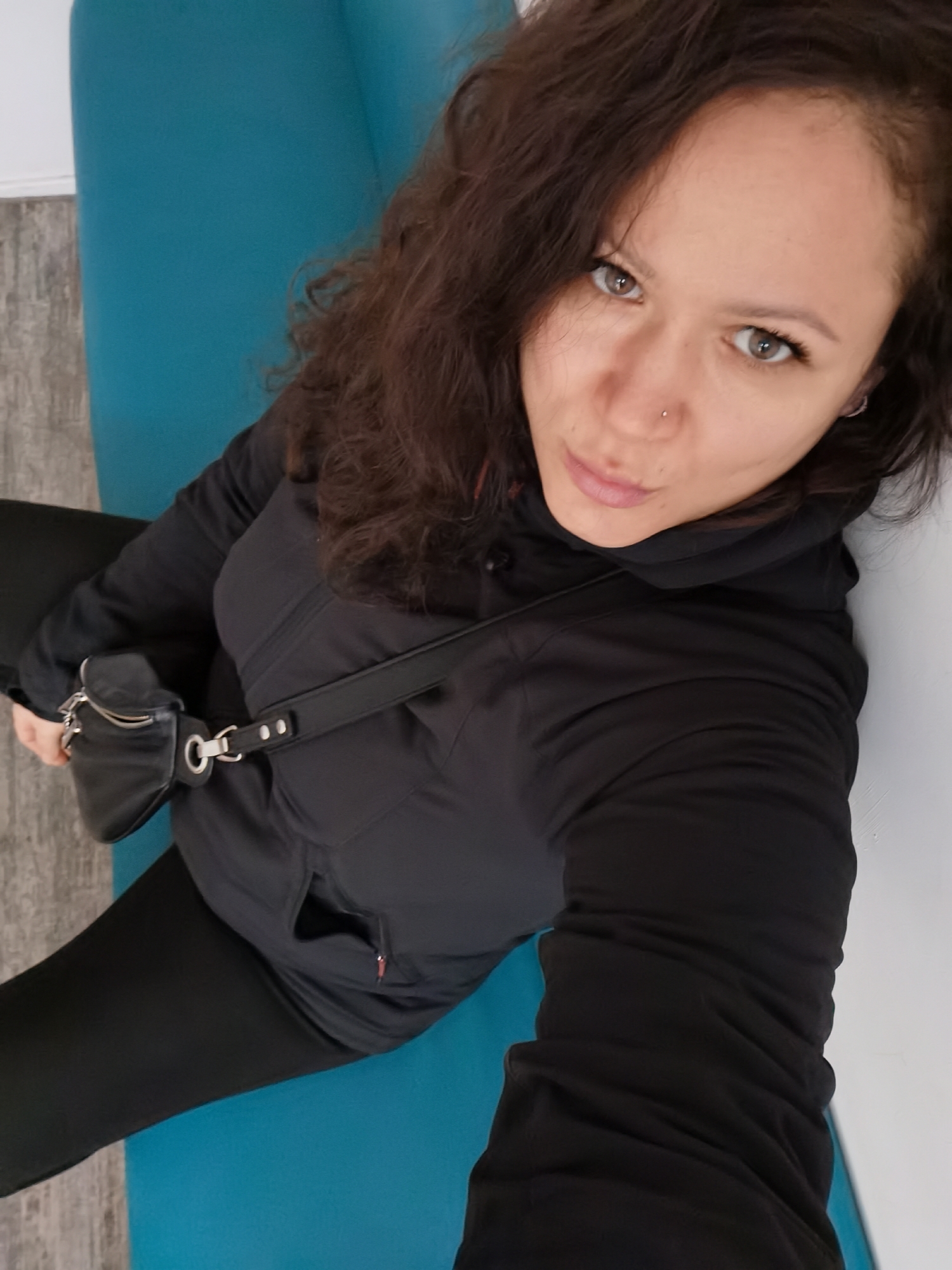 A woman with curly brown hair, a nose ring, and gray eyes is seated on a blue surface. She is wearing a black hooded jacket, black leggings, and a small black bag with a silver strap across her body. The setting appears to be indoors, with a light gray wall visible to the side. Based on the information provided, it is said that the location of the photo is in the city of Chaikovsky, Russia.