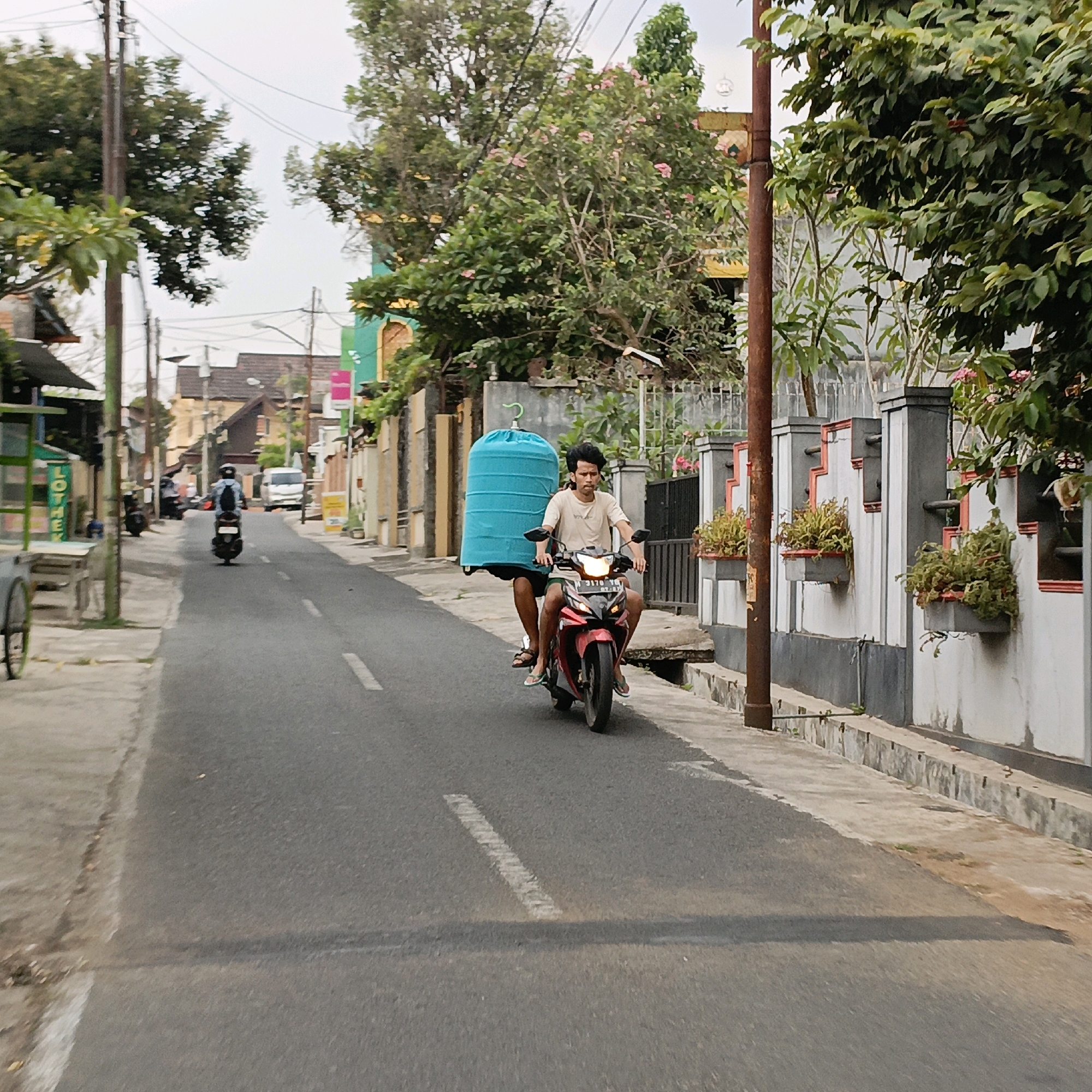 A man and a boy are riding on a red motorcycle on a street in Salatiga, Indonesia. The man is driving, and the boy is sitting behind him. They are both wearing light-colored shirts. A large, bright blue birdcage is strapped to the back of the motorcycle. The street is paved, and there are buildings and trees on either side. The buildings appear to be residential, with some shops visible. The sky is overcast. The image is taken from a slightly low angle, looking down the street.