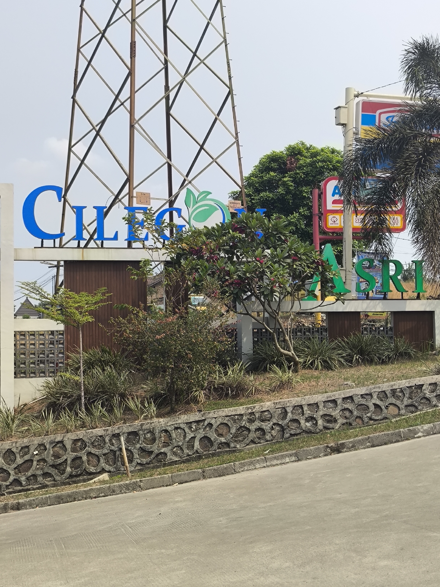 The image shows a large sign with blue and green lettering that reads "CILEGON ASRI". The sign is located in Cilegon, Indonesia, as indicated by the lettering. The sign is set against a backdrop of a tall lattice tower and trees. In the background, there are some buildings and a palm tree. The foreground shows a stone wall and a paved road. The weather appears to be overcast.