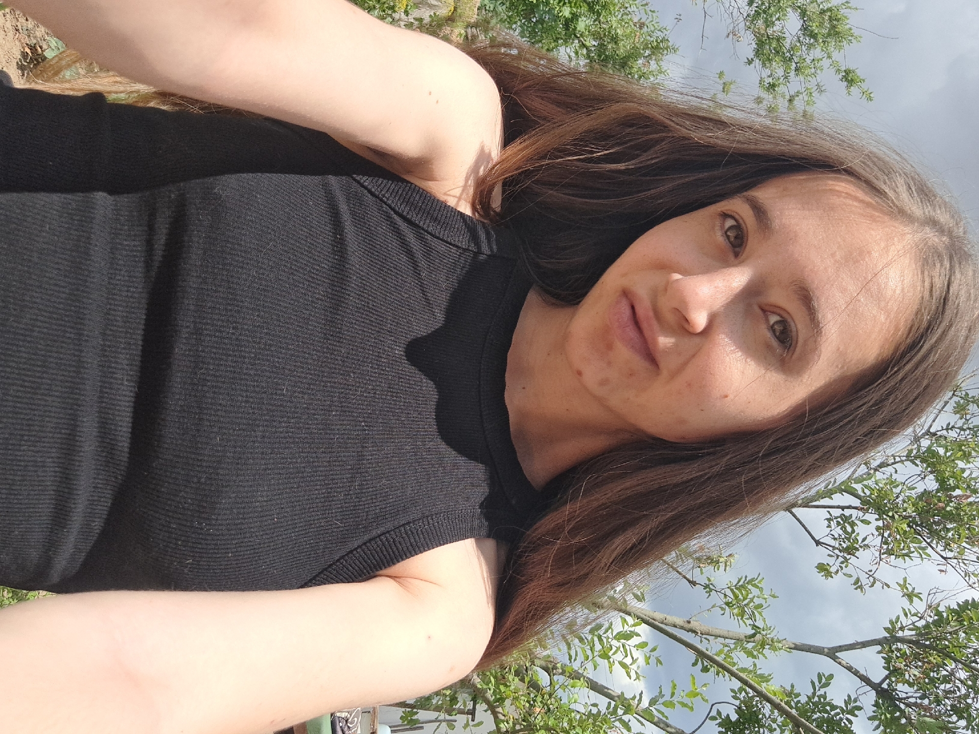 This is a close-up, selfie-style photograph of a young woman with long, brown hair. She is wearing a black ribbed tank top and is outdoors, with trees and a cloudy sky visible in the background. The scene appears to be set in a natural, possibly rural, environment. There is no indication of the city of Vypasne, Ukraine, in the image itself.
