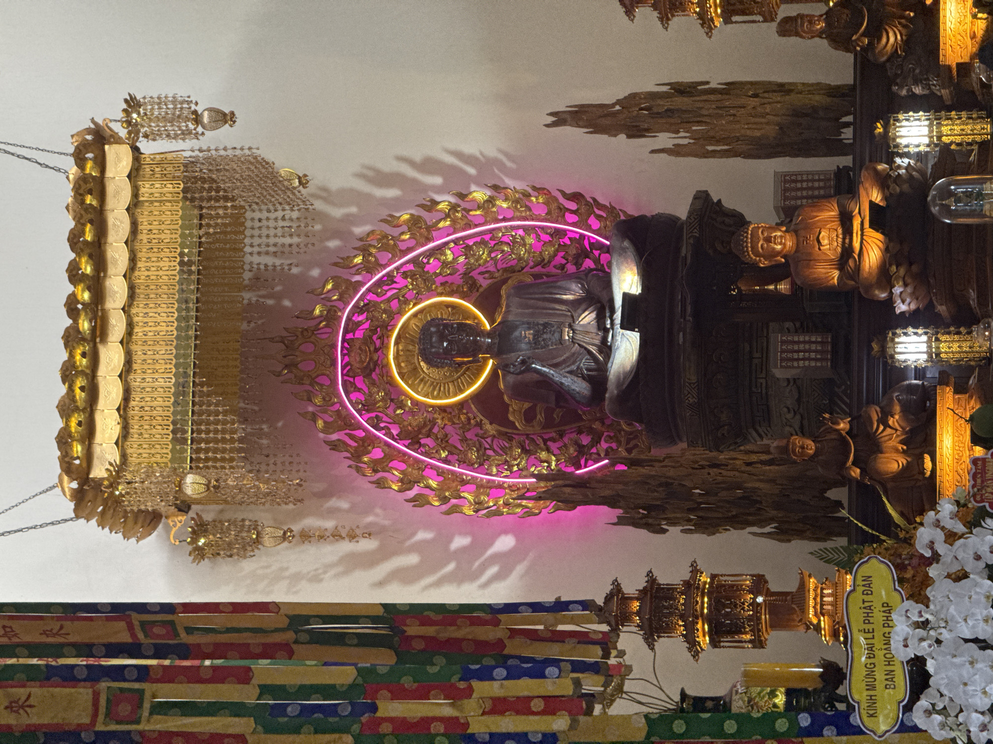 The image showcases the interior of a temple in Nha Trang Ward, Vietnam, focusing on a Buddhist altar. A central statue of the Buddha is illuminated by a pink and gold halo-shaped neon light. Ornate gold decorations adorn the altar, including a tiered structure hanging from the ceiling. Below the Buddha statue, there are additional statues and offering tables with lit candles. A sign in Vietnamese is visible, along with white orchids. The altar is covered with colorful, patterned cloths.