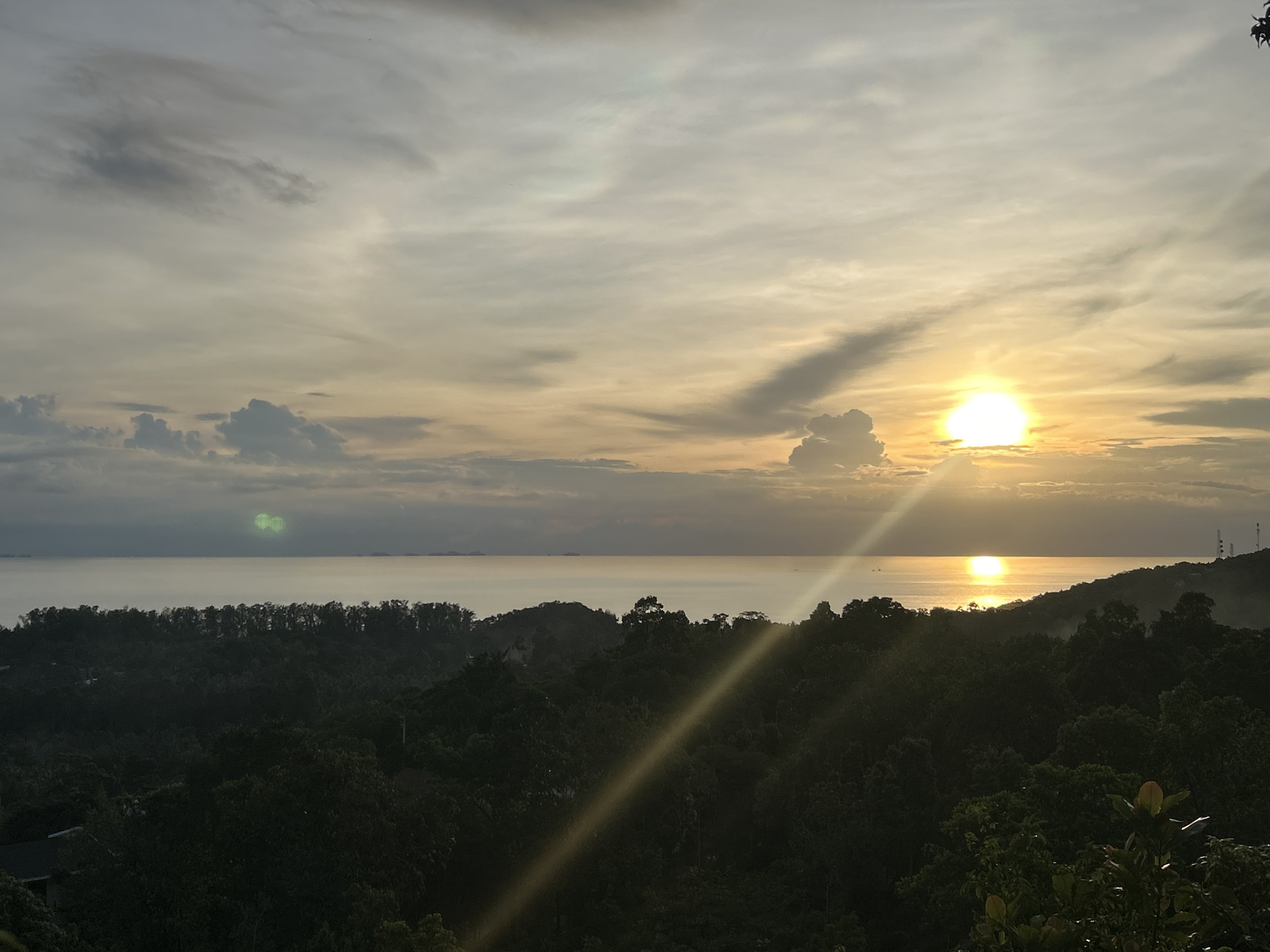 The image depicts a serene natural landscape at sunset, likely in the Koh Phangan subdistrict of Thailand. The foreground is dominated by a dense canopy of dark green trees, forming rolling hills that recede towards the sea. Sunlight filters through the foliage, creating dappled patterns. In the mid-ground, the calm, reflective surface of the ocean stretches horizontally across the frame. The horizon is marked by the distant silhouettes of islands or landmasses, and a few faint structures, possibly telecommunication towers, are visible on a hill in the far right. The sky above is a canvas of dramatic sunset colors, with the sun a brilliant, glowing orb positioned to the right of the center. Rays of light fan out from the sun, illuminating the clouds, which range from wispy streaks to heavier, darker formations. The overall atmosphere is tranquil and picturesque, characteristic of a tropical coastal setting during twilight.