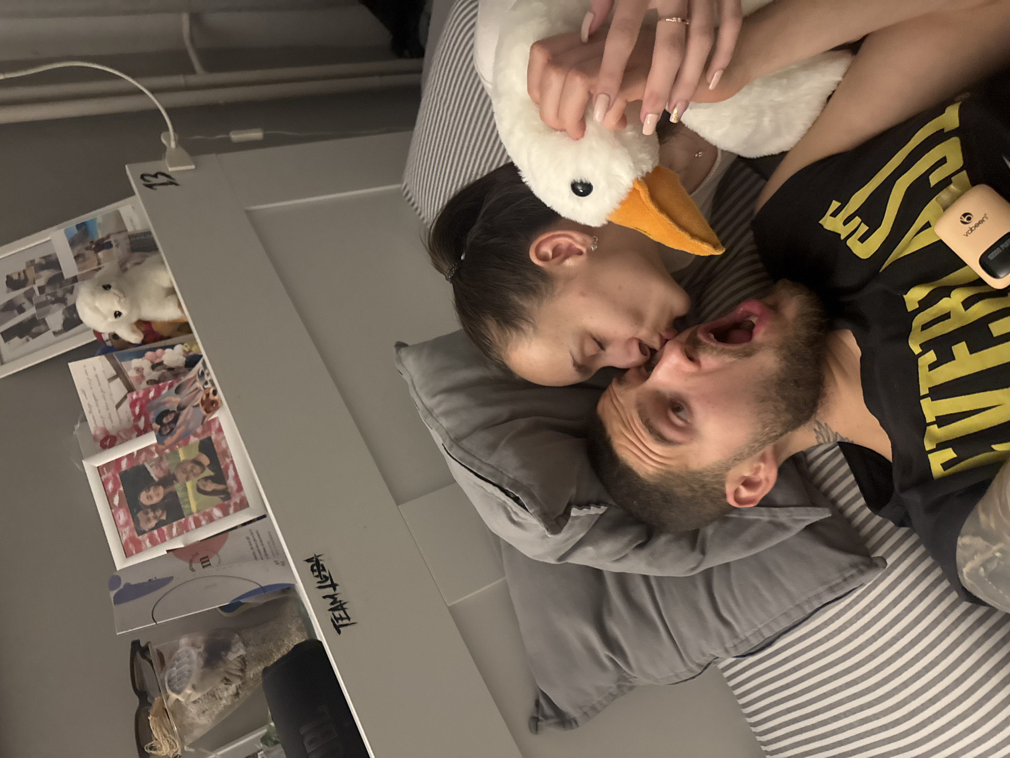 A couple is lying in bed. The woman is kissing the man. A white plush goose toy is positioned between them, held by the woman. The man has a beard and is wearing a black t-shirt with yellow lettering. A vaping device is visible near him. The setting appears to be a bedroom with a shelf visible on the side, displaying photos and other items. The text "TEAM LEBA" is written on the shelf. It is not possible to determine the location within the city of Sofia, Bulgaria based on the provided image.