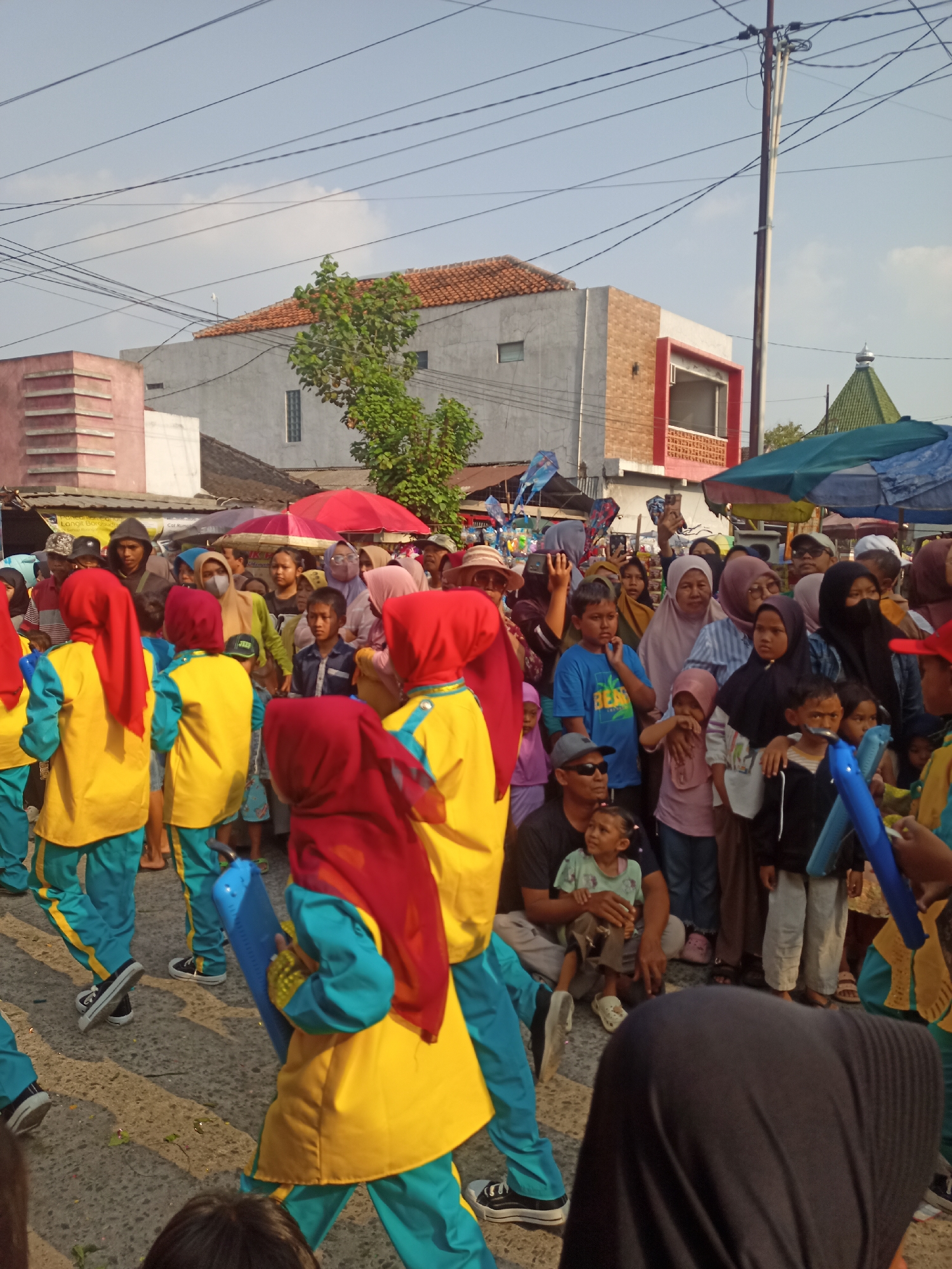 The image depicts an outdoor gathering of people in Saren, Indonesia, likely a street or public square. The scene is populated with a diverse crowd of adults and children, many of whom appear to be observing an event or procession. Several children in matching yellow and teal uniforms with red headscarves are prominent in the foreground, suggesting they are participants in a performance or parade. Some of these children are carrying blue objects, possibly props or instruments.
The environment includes buildings with varying architectural styles, some with red tile roofs and others with modern facades. Umbrellas and tents are visible, indicating a market or outdoor event. Electrical wires crisscross the sky, a common sight in urban and suburban areas. The overall atmosphere suggests a community event, possibly a festival or celebration. The lighting indicates it is daytime, with clear skies.