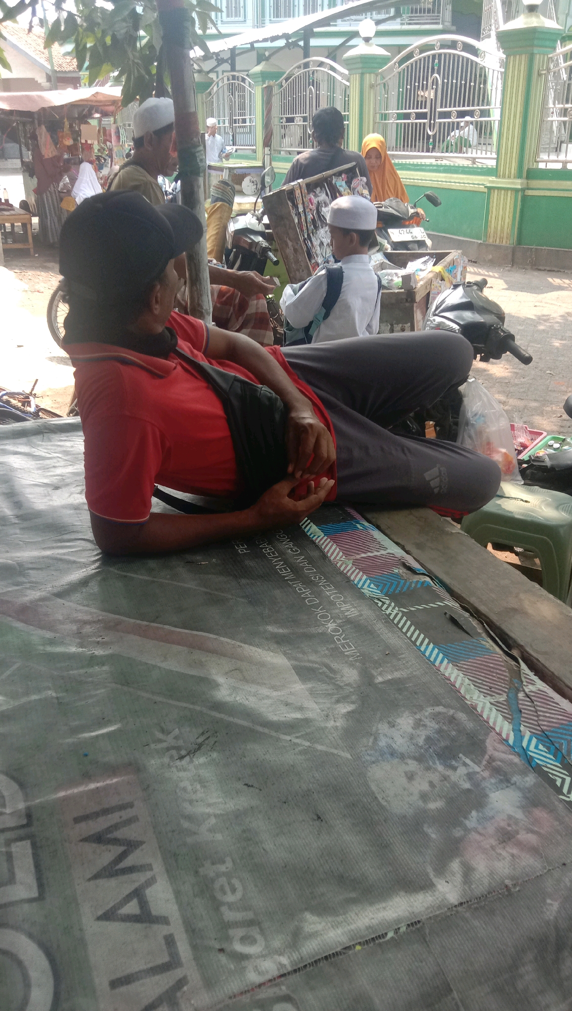 In front of a green and white building with a metal fence, several people are gathered outdoors. In the foreground, a person wearing a red shirt and dark pants is lying on a large, gray tarp that has some text printed on it. The person's left arm is bent, and their hand is resting on their lap. Behind them, a motorcycle is parked, with a wooden box attached to the back. Inside the box, there is a picture and some other items. A child wearing a white shirt and a white cap is standing next to the motorcycle, looking towards the box. Further in the background, other individuals are visible, some standing and some sitting. One person is wearing an orange headscarf. The environment appears to be a street in Ngempit, Indonesia, with market stalls visible to the left of the frame. The lighting suggests it is daytime.