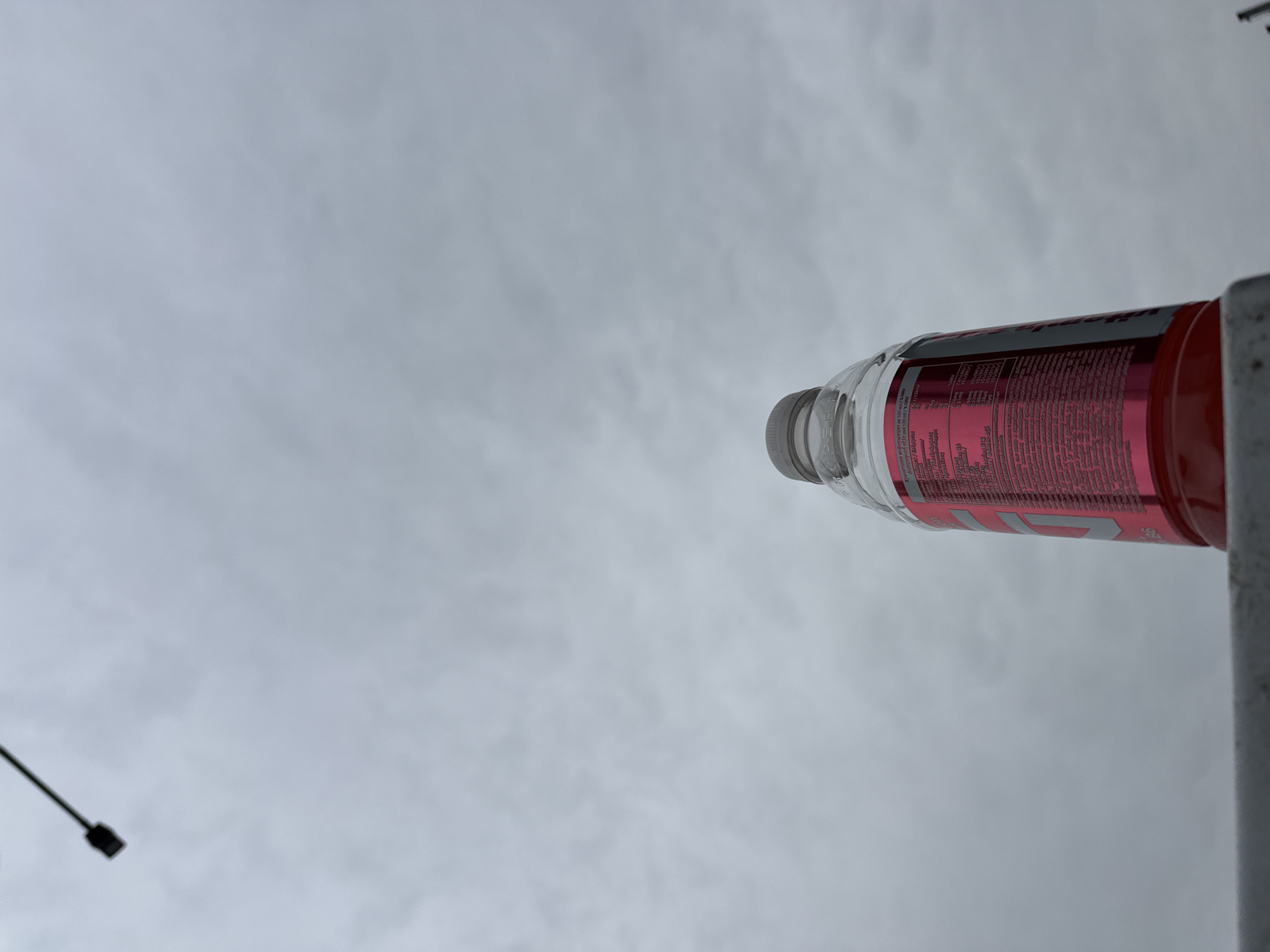 From a low angle, a clear plastic bottle with a grey cap and a reddish-pink liquid is visible against an overcast sky. The bottle's label contains nutritional information in multiple languages, including Greek. The bottle is partially obscured by a grey vertical bar, possibly a support structure or railing. In the lower left corner, a streetlight pole is seen. The location is likely in an urban area, but no specific details indicating Plovdiv, Bulgaria are present.