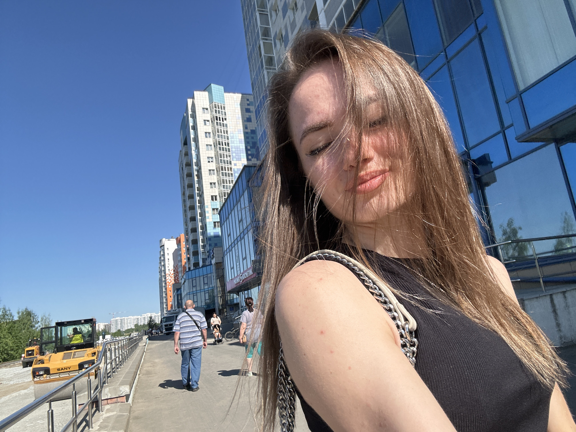 The image captures a close-up selfie of a young woman on a bright, sunny day in an urban environment. The woman is positioned on the right side of the frame, with her face partially shadowed by her long brown hair, which is blown by the breeze. She wears a black sleeveless top and has a bag with a prominent chain strap over her shoulder. Her eyes are partially closed or in shadow, and her lips are slightly pursed.
The background reveals a bustling street scene with several modern multi-story buildings. Some buildings feature reflective blue glass facades, while others have white and blue paneling, with one exhibiting orange and white checkered patterns. A clear, deep blue sky dominates the upper left portion of the image.
On the left side of the frame, there's evidence of road construction or maintenance, with a yellow SANY road roller clearly visible, and another similar vehicle further in the distance. Several people are walking along a paved sidewalk that has a metal railing. A man in a striped shirt and blue jeans walks away from the camera, and another person pushing what appears to be a stroller is visible further back. Green trees and bushes line the construction area on the far left. The overall impression is one of a vibrant city street on a clear, sunny day, likely midday or early afternoon, given the strong shadows. Faint Cyrillic text, possibly "МАГНИТ КОСМЕТИК" (Magnit Kosmetik), can be seen on a building in the background.