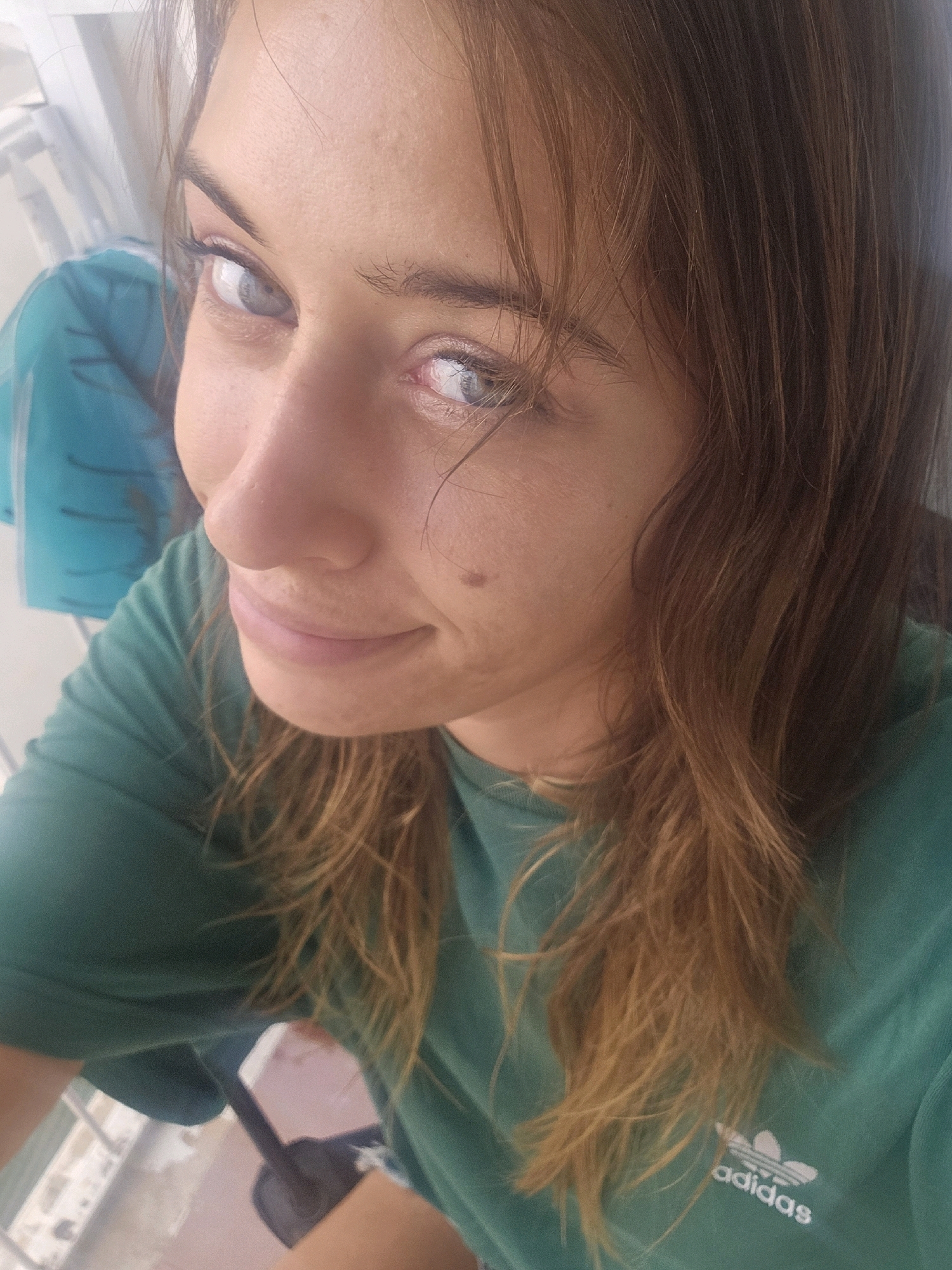 A close-up, high-angle shot captures a young woman with long, light brown hair and light blue eyes. She is wearing a green t-shirt with an Adidas logo. She is looking towards the camera with a slight smile. The background is blurred, but appears to be an outdoor setting with a balcony or railing and some blue fabric. The scene is likely in Palma de Mallorca, Spain, as indicated by the presence of an Adidas logo, which is a globally recognized brand, and the specific mention of the city and country in the prompt, suggesting a contextual association.