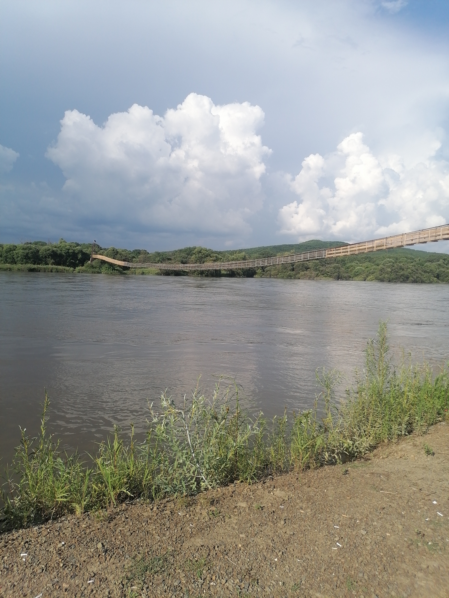 The image shows a wide, murky river flowing through a vegetated landscape. In the background, a long, wooden suspension bridge spans the river, connecting two tree-covered banks. The bridge appears to be an older structure with a weathered wooden walkway and railings. The sky above is partly cloudy, with large white cumulus clouds contrasting with patches of blue. The foreground of the image is a dirt and gravel embankment with some sparse green vegetation growing along the water's edge. The scene is calm and natural, suggesting an outdoor recreational area or a natural landscape. The location is identified as Novopokrovka, Russia.