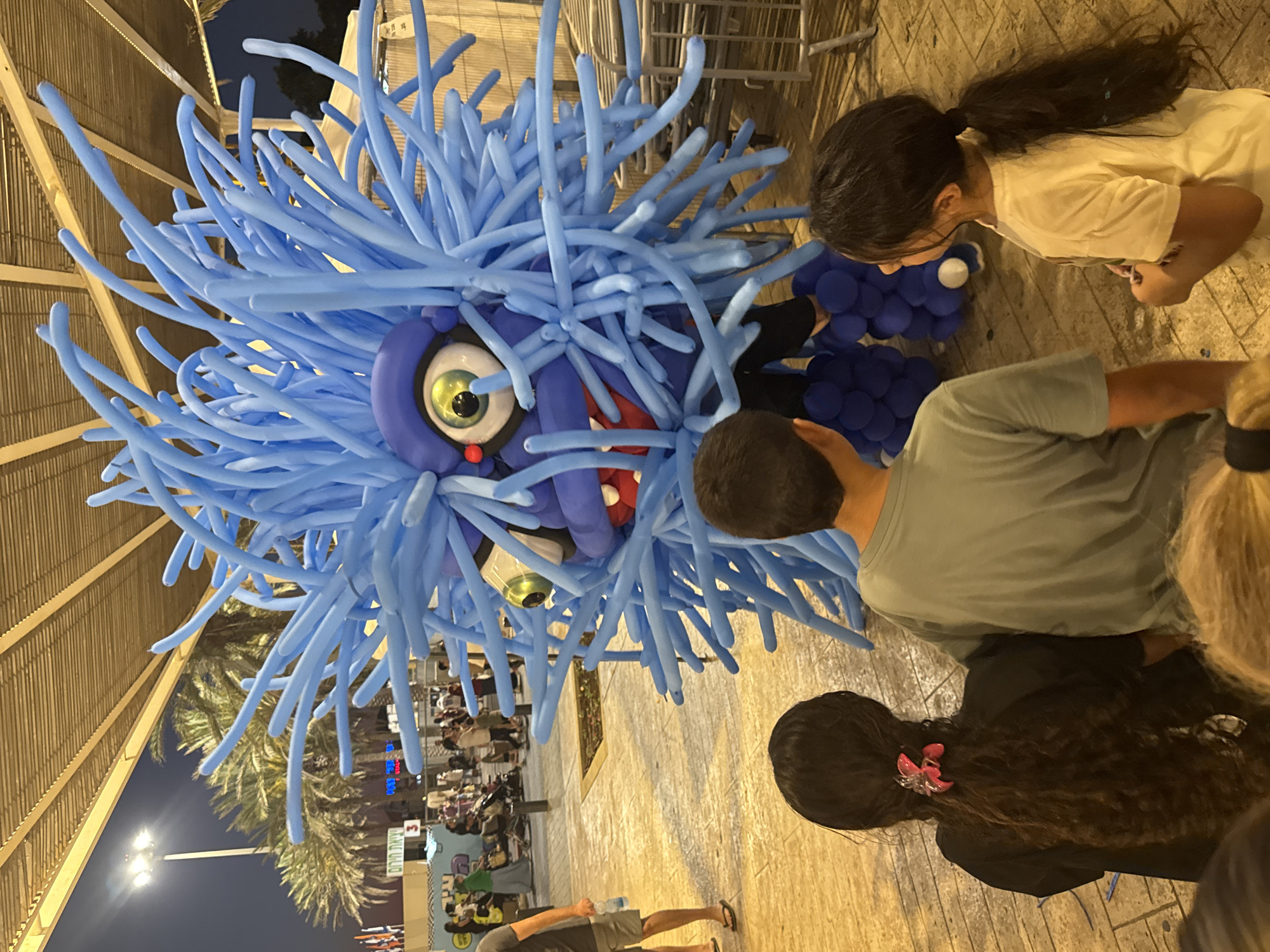 At night, a large blue balloon creature with numerous tendrils stands in an outdoor plaza in Netanya, Israel. The creature has one large, green, bulging eye with a red pupil, and a wide mouth with sharp, red teeth. It is surrounded by a crowd of people, including three children in the foreground looking at it. The plaza appears to be paved with cobblestones, and palm trees are visible in the background. There are also tents and temporary structures, suggesting a fair or festival. The lighting is artificial, casting a warm glow on the scene.