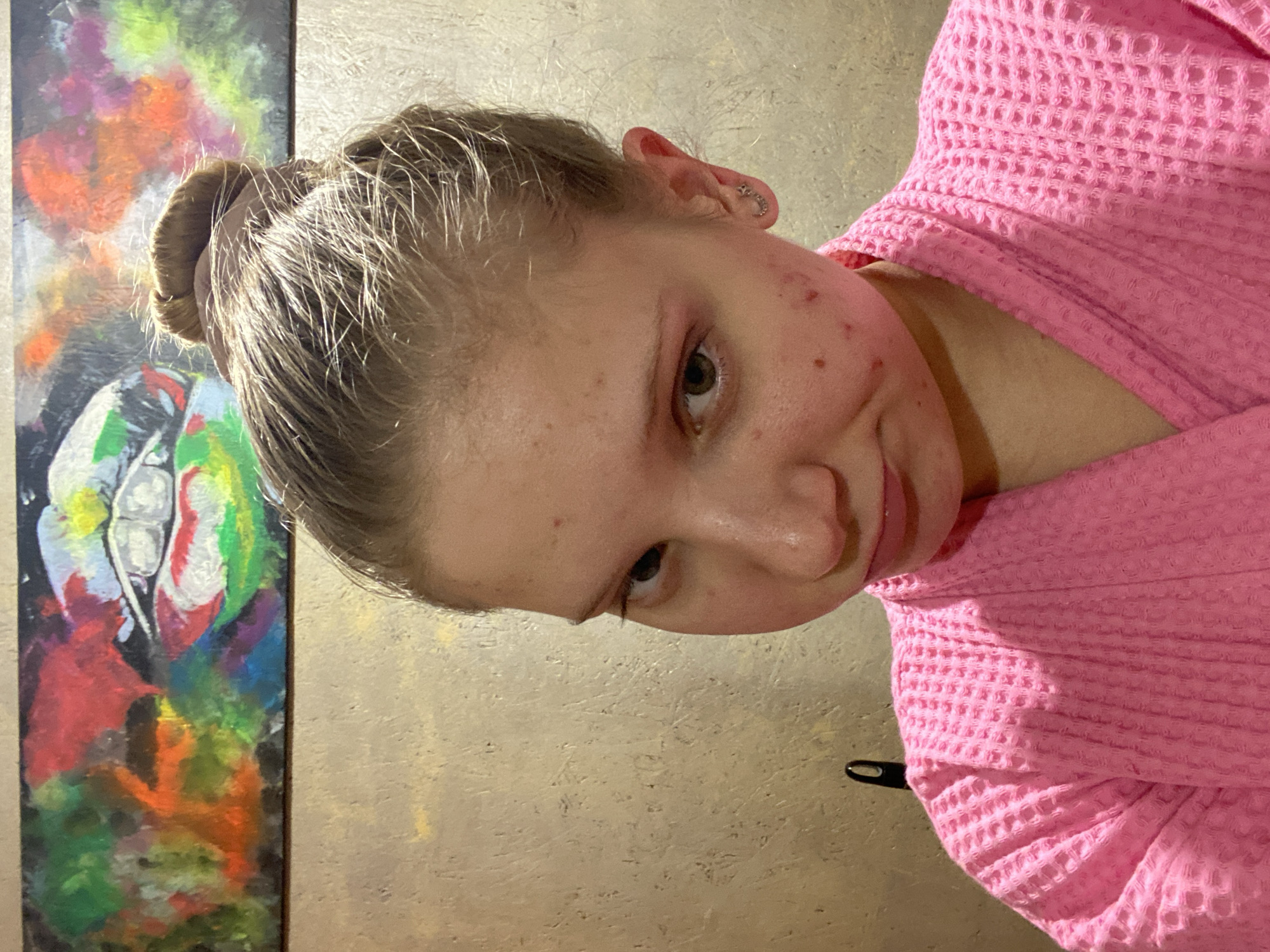 A fair-skinned young woman with light brown hair styled in a bun and wearing a pink waffle-weave robe is shown in an indoor setting. She has blemishes on her face and a piercing in her ear. The background consists of a light tan wall with a dark-framed painting featuring a colorful abstract design to the left. The image appears to be taken indoors, likely in a personal residence. The location is the city of Chaikovsky, Russia.