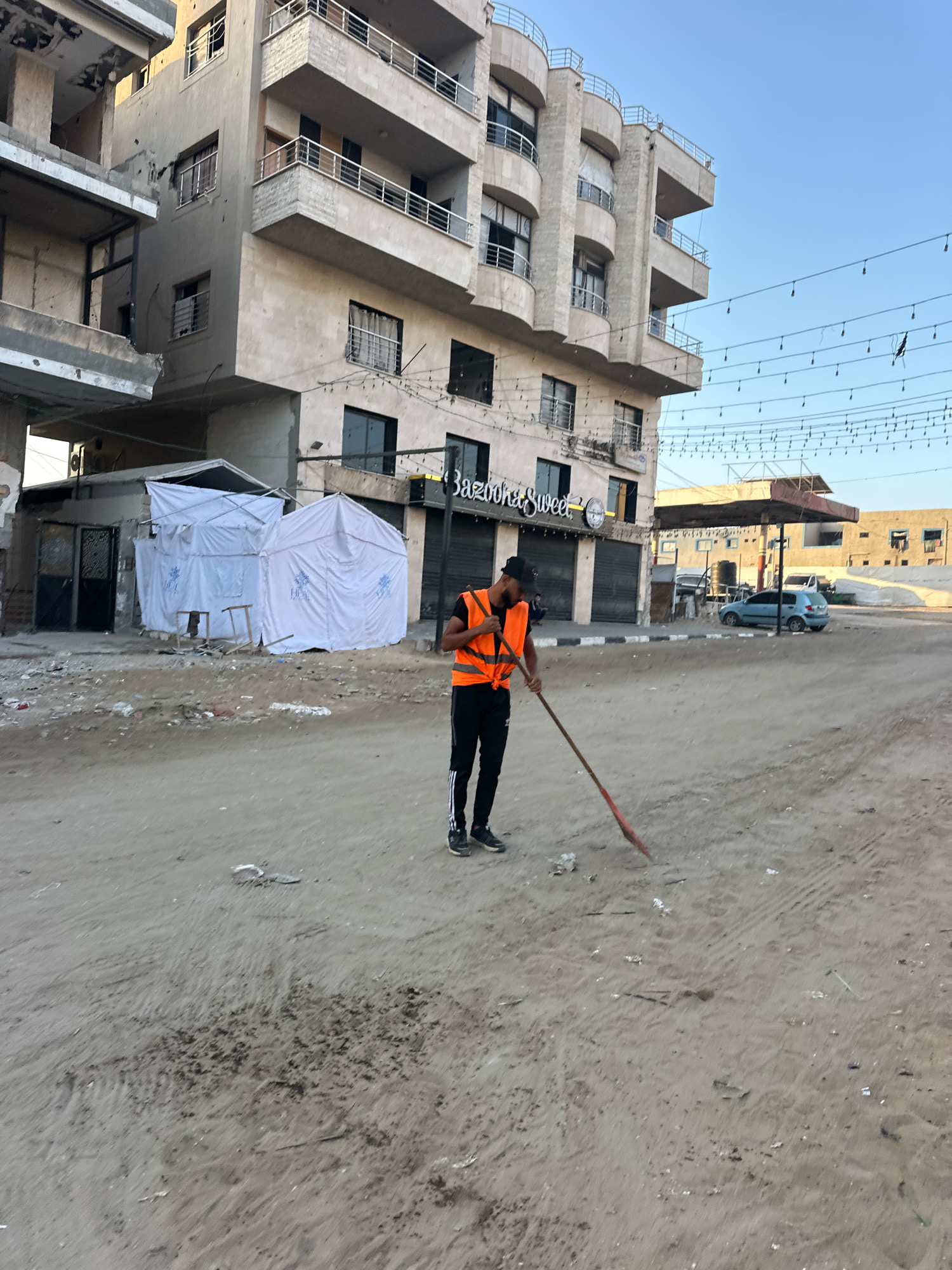 In Gaza City, Palestinian Territories, a young man wearing an orange safety vest and black pants with white stripes is sweeping a dirt road with a red-tipped broom. The street appears to be dusty and littered with debris. Behind him, a multi-story building with balconies and broken windows stands next to two white tents. Another building with a sign that reads "Bazooka Sweet" is visible further down the street. A light blue car is parked near a gas station in the background. The sky above is clear and blue.