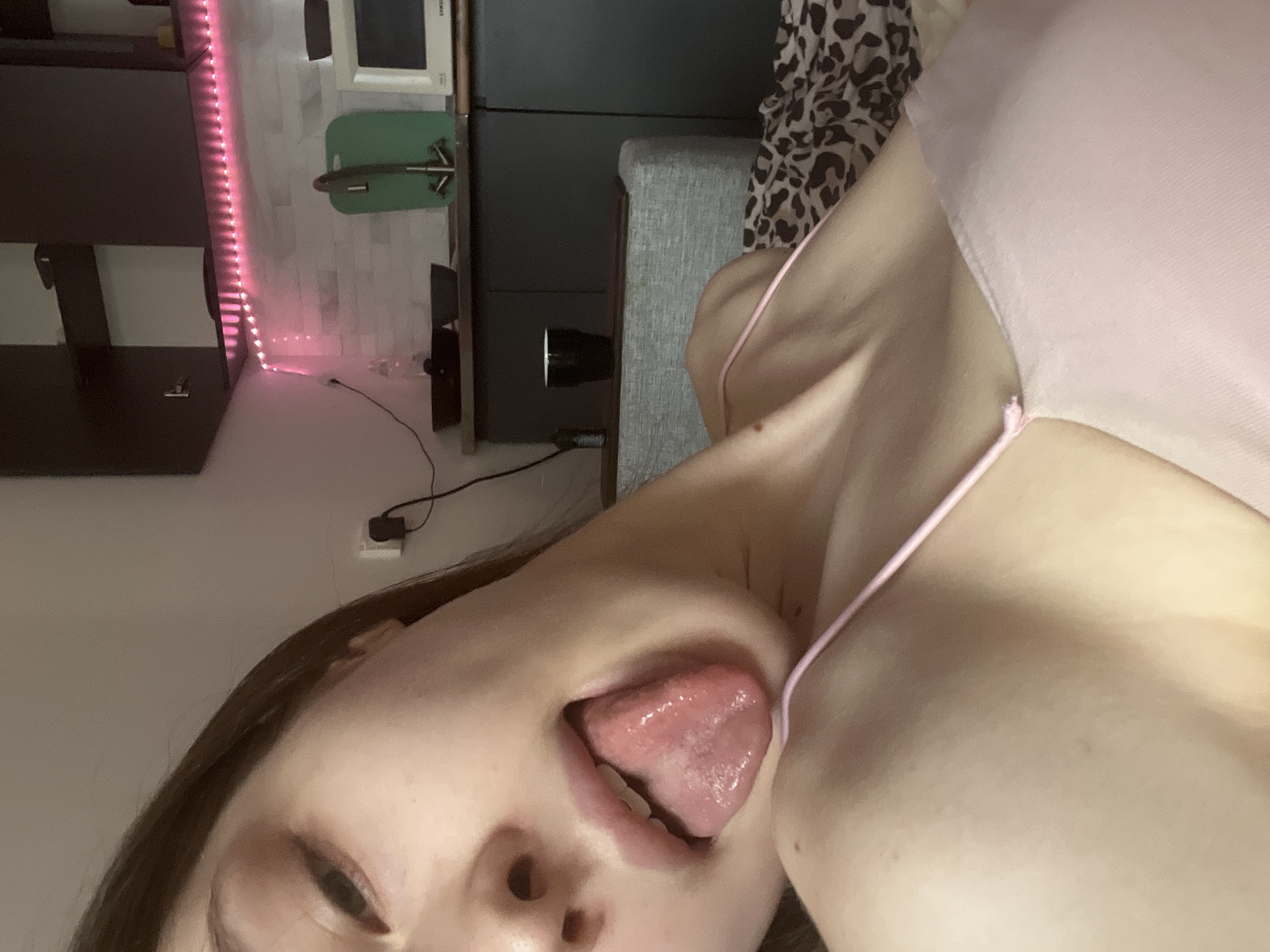 A close-up, angled selfie photograph features a woman with her mouth open, extending her tongue. She is wearing a pink camisole. The background includes a black cabinet or shelving unit with pink LED strip lights and a gray upholstered item which could be a headboard. There is a white object and a green object on top of the black cabinet. No evidence suggests the location is Saint Petersburg, Russia.