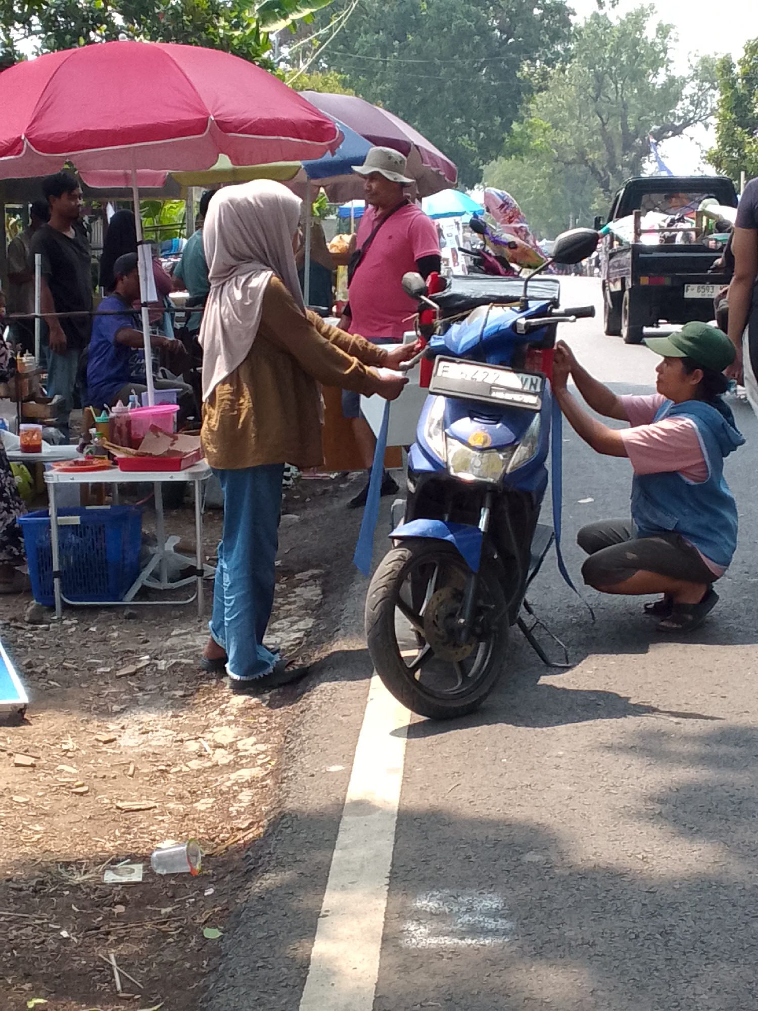 The image depicts an outdoor scene, likely a street market, in Mandalawangi, Indonesia. In the foreground, a blue motorcycle is being attended to by two individuals. A person wearing a light brown hijab and blue jeans is standing to the left, while another person wearing a green cap and a denim vest over a pink shirt is squatting to the right, attaching a blue ribbon to the motorcycle. The motorcycle has a license plate displaying "F 6422 VN". To the left of the motorcycle, a small table laden with food items, sauces, and containers is visible. Blue plastic baskets are placed beneath the table. In the background, several people are gathered under red and purple umbrellas, suggesting a commercial or social gathering. A black pickup truck with items in its bed is parked further down the road. The ground is a mixture of dirt and asphalt, with a white line marking the edge of the road. Trees and foliage are visible in the background, indicating an outdoor environment.
