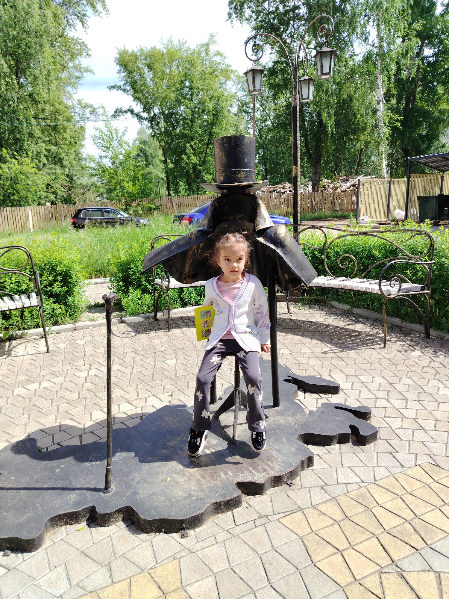 A young girl with dark, curly hair sits centered on a unique metal sculpture in an outdoor park in Chaikovsky, Russia. The sculpture features a dark, silhouette-like figure wearing a top hat and a flowing coat, with a walking cane leaning against it. The girl, appearing to be a toddler or preschooler, is seated on the stool element of the sculpture. She wears a white cardigan with purple embroidered details over a light pink top, grey pants with white butterfly or leaf patterns, and black shoes. In her hands, she holds a bright yellow book with small illustrations and what appears to be Cyrillic text, partially visible as "У" or "Ч". Her expression is neutral as she looks directly at the camera.
The scene is brightly lit by sunshine, indicating midday or early afternoon, with distinct shadows cast on the ground. The paving beneath the sculpture is composed of light-colored, geometrically shaped tiles. To the right, an ornate wrought iron street lamp with three lanterns and decorative scrollwork rises. Two additional wrought iron benches, whose seats are designed to resemble piano keys, are visible on either side of the main sculpture.
The background features lush green trees and dense bushes, suggesting a vibrant natural setting. A light wooden fence runs along the perimeter behind the greenery, with parts of a black SUV and a blue car partially visible beyond it. The clear blue sky is partially visible through the tree canopy. The overall setting is a pleasant public space, likely a park or garden, with artistic elements that subtly hint at a musical or classical theme, appropriate for the town's namesake.