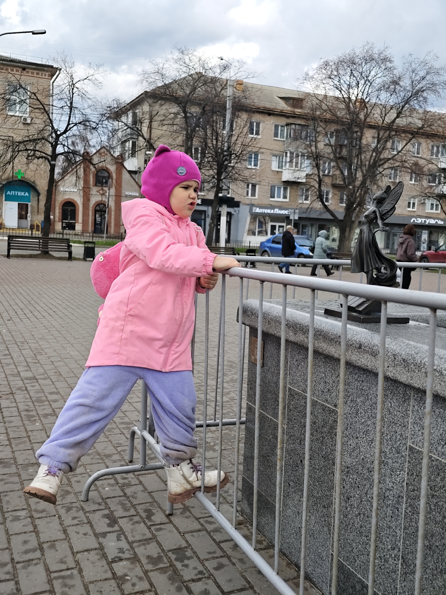 A young girl, estimated to be between three and five years old, is depicted in an outdoor public space in Chaikovsky, Russia, during an overcast daytime. She is dressed in a bright pink winter hat with earflaps, a pink jacket, and light purple fleece pants, with white boots on her feet and a small pink backpack visible on her back. The girl stands with one foot on the lower bar of a grey metal barrier, leaning against the top bar, and appears to be looking with a focused expression towards a dark bronze statue of a winged figure (possibly an angel or fairy) on a granite pedestal.
The setting appears to be a town square or pedestrian area with grey rectangular pavers. In the background, several adult pedestrians are walking. Behind them, various multi-story buildings with Eastern European architectural styles are visible. Bare trees, indicative of late autumn, winter, or early spring, are scattered throughout the scene. Visible Cyrillic text on storefronts includes "АПТЕКА" (Pharmacy), "КОФЕЙНИ" (Coffee Shops), "АПТЕКА ПЛЮС" (Pharmacy Plus), and "ЛЭТУАЛЬ" (L'Etoile, a cosmetics store). The overall atmosphere is cool and cloudy.