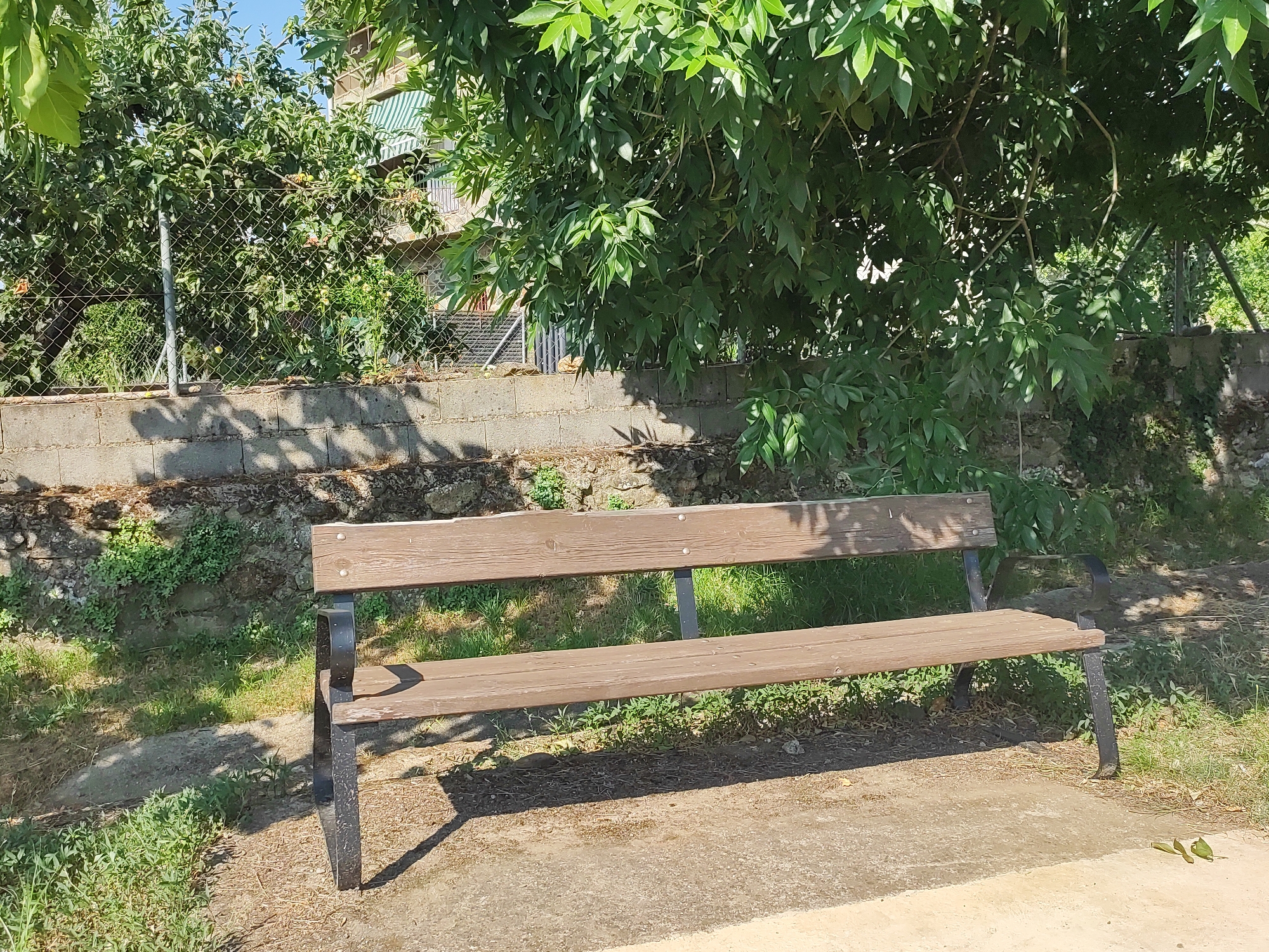 A wooden park bench with black metal legs sits on a dirt path in front of a low stone wall. Behind the wall, a chain-link fence encloses trees and foliage. A building with green and white awnings is visible through the trees. The scene is outdoors in daylight.