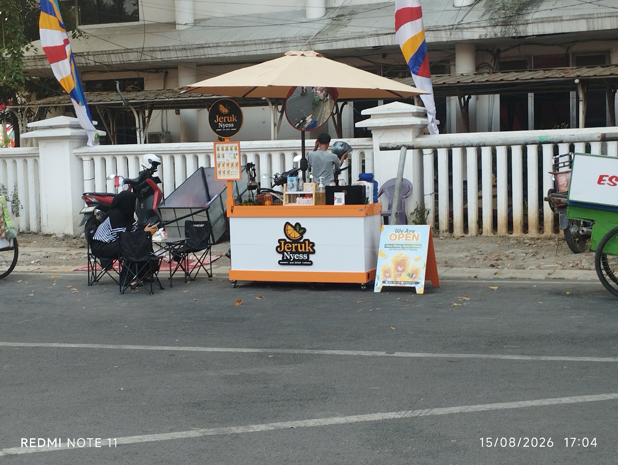 The image depicts an outdoor scene in Karawang, Indonesia, with a focus on a small juice stand. A young person, possibly a vendor, is seen behind the counter of an orange and white cart, wearing a grey t-shirt and facing away from the camera. The cart has an umbrella providing shade and is adorned with a circular logo that reads "Jeruk Nyess" and "est 2021". To the right of the cart is a sign that says "We Are OPEN" with images of juice glasses. In the background, a white fence runs along the property line, and a building with multiple windows is visible. A few parked motorcycles and chairs are scattered around the scene. Two flags, one red, white, and blue, and another with white, yellow, and blue stripes, are displayed on poles. In the foreground, the asphalt road is visible, with white lane markings. The bottom of the image shows the text "REDMI NOTE 11" and the date "15/08/2026 17:04".
