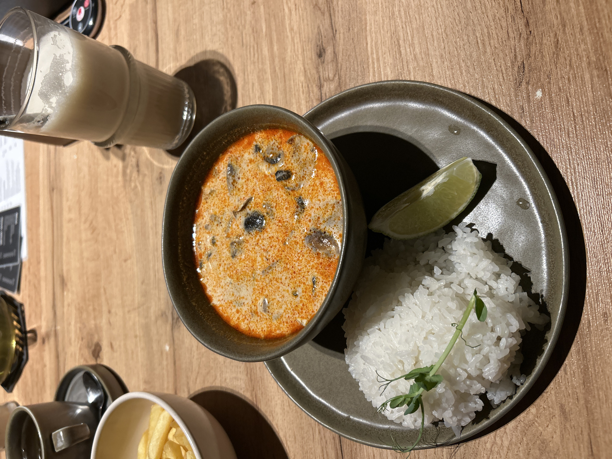 This overhead shot captures a meal laid out on a light-colored wooden table. The central subject is a dark gray ceramic bowl filled with a creamy, orange-colored soup, featuring visible dark pieces, possibly mushrooms or meat. Beside the soup, on a matching dark gray plate, is a generous portion of white, fluffy rice, garnished with a delicate green sprout. A fresh lime wedge rests on the plate next to the rice.
In the upper left quadrant, a tall, clear glass contains a light, opaque beverage with froth on top, suggesting a milkshake or a similar creamy drink. Partially visible in the lower left are a small dark gray mug and a light-colored bowl containing French fries.
The scene depicts a meal being served in what appears to be a restaurant or cafe setting. Prominent shadows cast by the dishes and glass suggest direct overhead lighting. The wooden table exhibits a natural grain. Blurred text, likely from a menu or bill, is faintly visible in the background near the glass. No individuals are present in the image. The meal is presented neatly, implying a dining experience.
