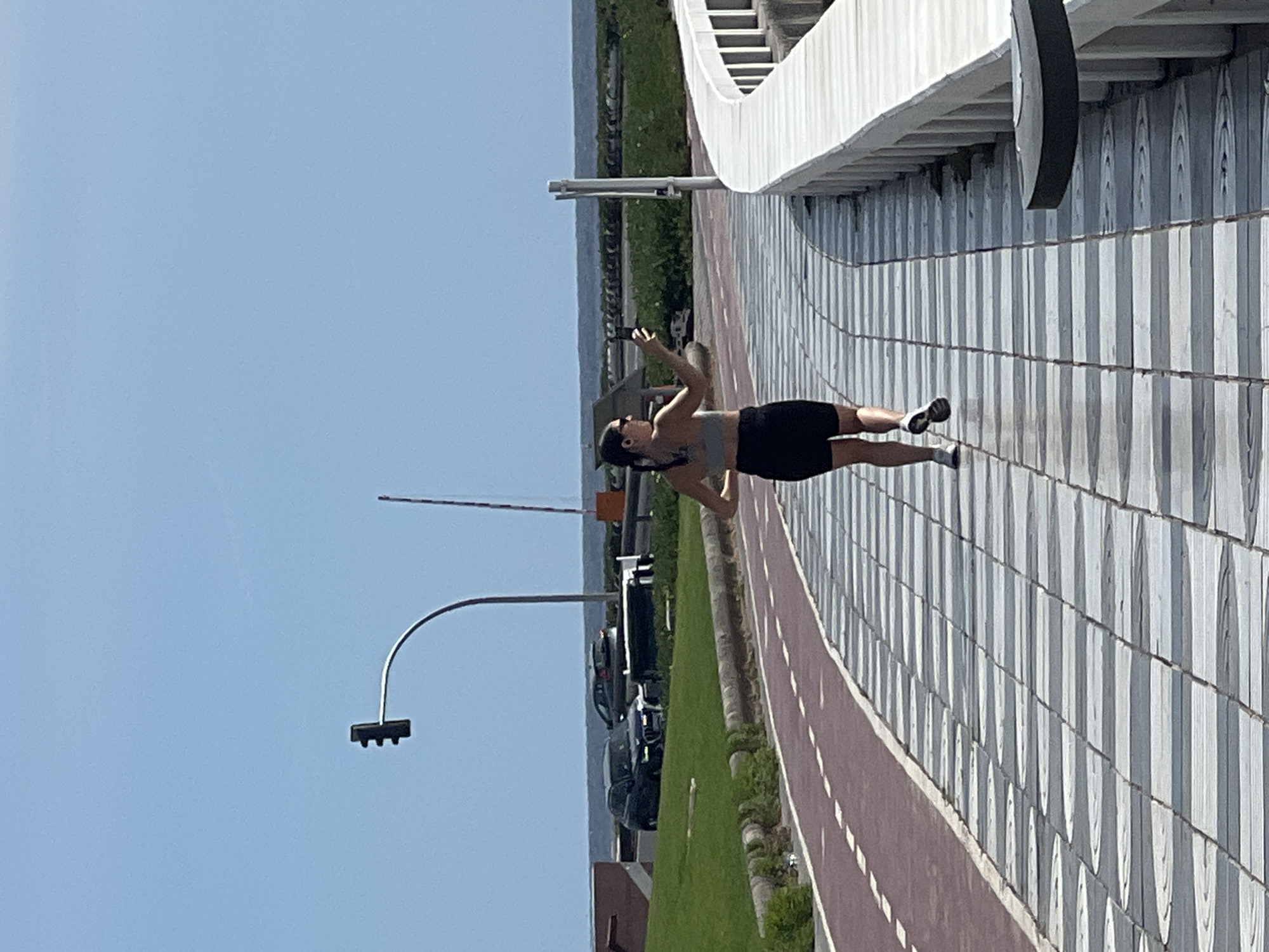 The image depicts a woman standing on a sidewalk next to a curved structure with white railing on a sunny day. She is wearing a sports bra, dark shorts, and sunglasses. She is holding her phone up, likely taking a picture or video. A paved pedestrian path runs alongside the sidewalk, adjacent to a green area. Beyond that is a road with cars and street lights.
This image likely shows the area near the Auditorio Alfredo Kraus in Las Palmas, Spain, based on the distinctive design of the structure with white railing.