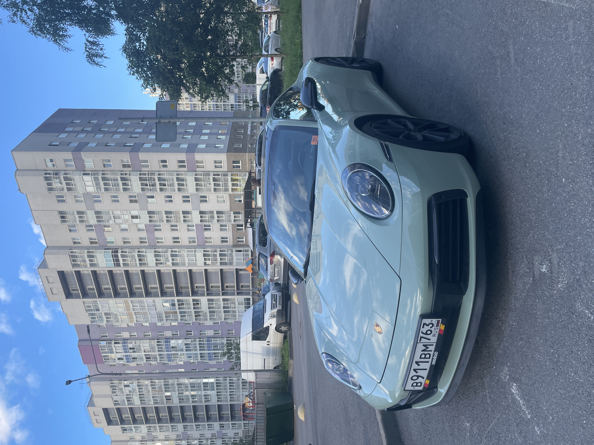The image shows a light green Porsche 911 parked on an asphalt road in front of a tall, modern apartment building. The building is grey and white with numerous windows and balconies, some of which are closed. A tree with green leaves partially obscures the top of the building. The sky is blue with scattered white clouds.
In the background, several other vehicles are visible, including a white van and other cars parked along the roadside. A yellow lamppost is mounted on the side of the building. There is also a green trash bin and some playground equipment visible near the base of the building.
The license plate of the Porsche is visible and reads "B911BM 763". Below this, "RUS" is printed, along with a small flag of Russia. Another small plate below that appears to say "josgoodrich".
The image is taken from a low angle, looking upwards towards the car and the building. The lighting suggests it is daytime, likely late morning or early afternoon, with clear weather. The scene appears to be in a residential area. There are no people clearly visible in the image. The overall mood is neutral and observational.