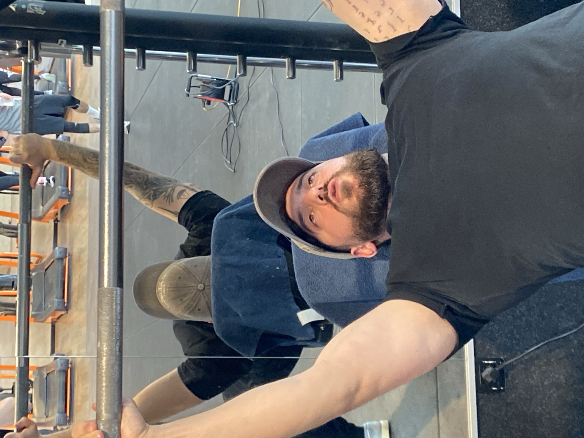 An adult male is depicted performing a bench press exercise within a gym in Hœnheim, France. The subject, exhibiting a short beard and wearing a grey baseball cap reversed along with a black t-shirt, is positioned supine on a dark padded bench. He holds an unweighted metallic barbell with an overhand grip, arms extended upwards. His left arm is visibly muscular, while his right forearm and bicep display extensive tattoos, including script and graphic patterns. A blue towel is draped over the top of the bench, beneath his head. Directly behind the subject, a large mirror reflects his back, the reverse of his cap, and the towel. This reflection also reveals several orange-accented treadmills in the background and partially visible figures of other individuals exercising. To the right of the subject, a metallic upright with multiple adjustment pegs, consistent with a power rack or spotter arm assembly, is present. The floor is a dark grey speckled material. Visible on the floor near the rack are various electrical cables and a black and red electrical device. A power outlet is visible on the dark base of the wall to the lower left.