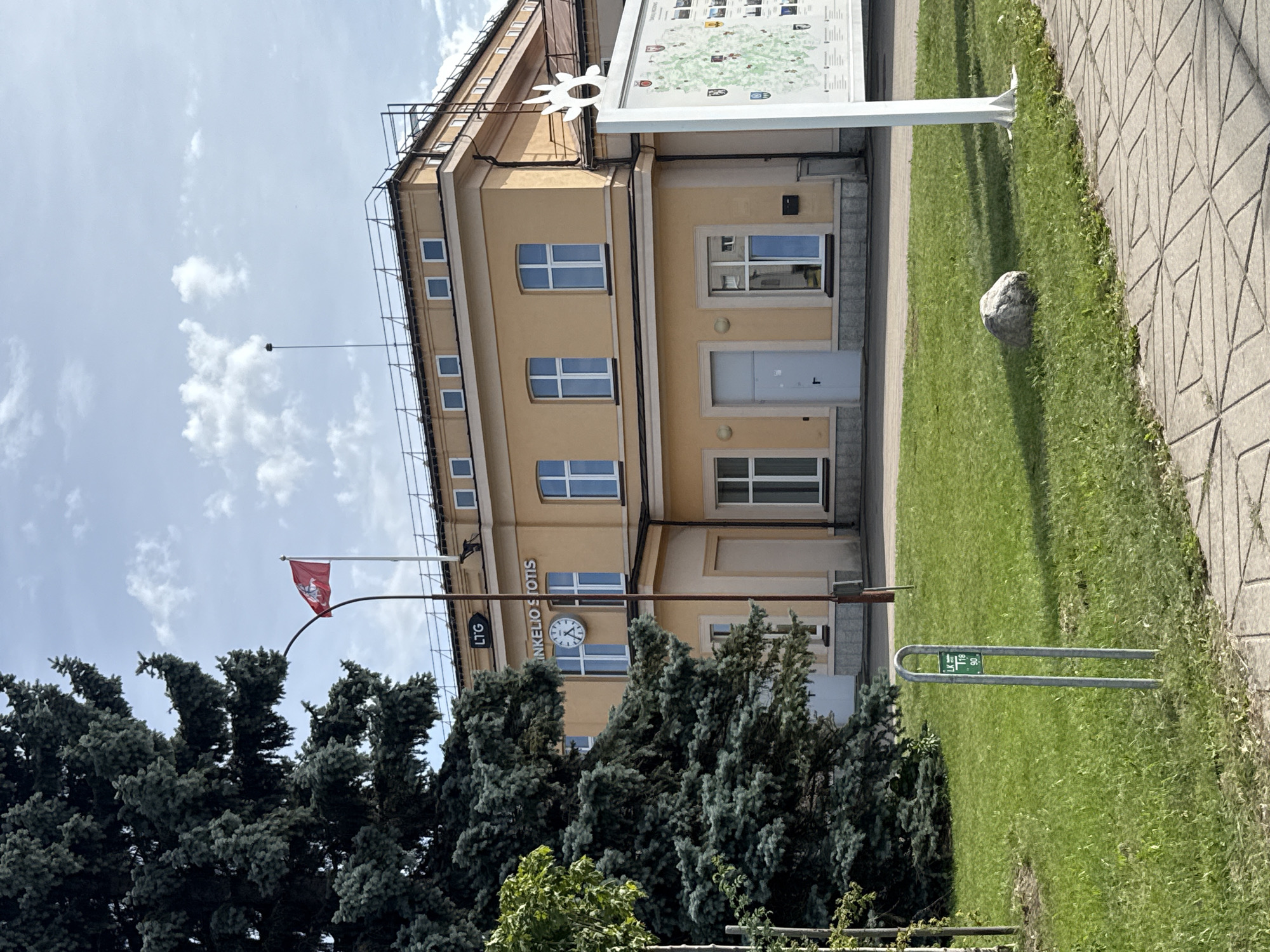 The image captures the exterior of the Šiauliai railway station in Šiauliai, Lithuania. A tall, dark green coniferous tree dominates the left side of the frame, partially obscuring the building. The building itself is a two-story structure painted in a pale yellow hue. A red flag with a white emblem flies from a flagpole atop the building. Above the entrance, the words "LTG" and "INKELIO STOTIS" are visible, indicating it is a railway station. A large clock is mounted on the facade. Several windows with white frames are visible on both floors. To the right of the building, a white information board with a map and various emblems stands on a pole. The foreground features a well-maintained green lawn with a silver bicycle rack, a gray stone, and a paved walkway. The sky is blue with scattered white clouds. No people are present in the image.