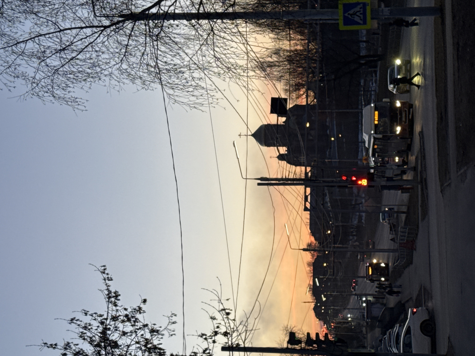 The image captures a street scene in Berezniki, Russia, at dusk. The sky transitions from light blue to orange near the horizon. Silhouetted against this backdrop is the Church of the Intercession of the Holy Virgin, identifiable by its dome and cross. Utility poles and wires crisscross the sky. On the right side of the frame, a road with pedestrian crossing is visible. A bus and cars are present, with their headlights illuminated. Pedestrians are visible on the sidewalk, some crossing the road. A traffic light is illuminated red. The environment appears to be urban, with streetlights and buildings lining the road. Bare branches of trees are visible along the top and bottom edges of the frame.
