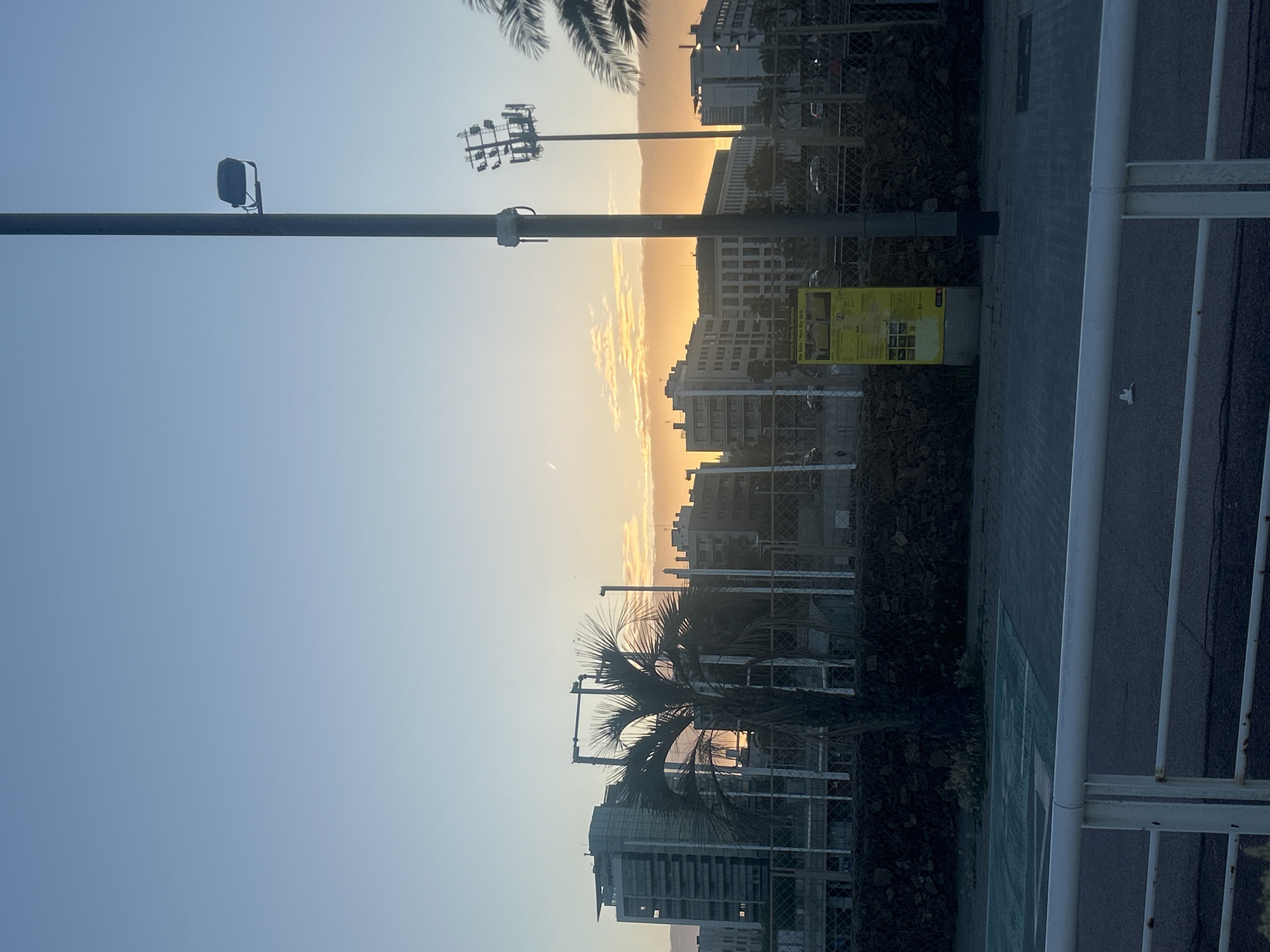 The image shows a view of the Barcelona cityscape at sunset. In the foreground, there is a metallic fence and a lamppost. The middle ground is occupied by buildings and palm trees, silhouetted against the setting sun. The sky ranges from light blue to golden orange. No people are visible. The image appears to have been taken on or near a street, overlooking the city. There are signs of rock or concrete walls or structures behind the fence.