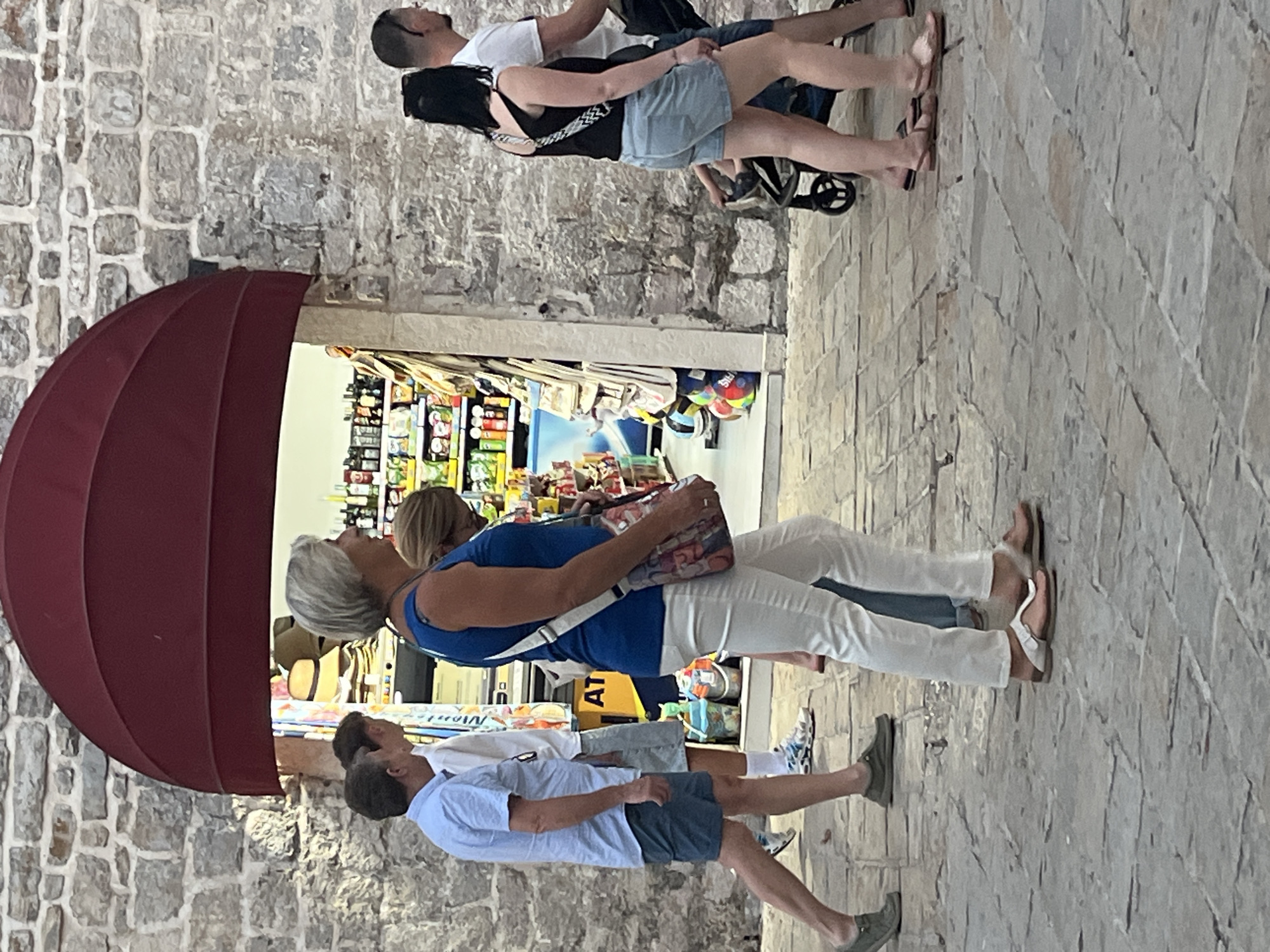 The image captures a street scene in the old town of Budva, Montenegro, near the Mediterranean Sea. The architecture features a weathered stone wall, characteristic of ancient European towns. A deep red awning protrudes over an archway, leading to a shop interior stocked with various goods, including shelves of bottles and what appear to be hats on display.
In the foreground, several people are walking along a cobblestone path. On the left, a man in a light blue short-sleeved shirt and dark blue shorts walks away from the camera. To his right, a woman in a blue tank top and white pants is also walking, carrying a colorful bag over her shoulder. Behind her, another person is partially visible. On the right side of the image, a woman in a black tank top and denim shorts walks with a stroller. A man in a white t-shirt walks next to her, and another person, possibly a child, is seated in the stroller. The people are casually dressed, suggesting a warm, pleasant day. The overall atmosphere is that of a lively, tourist-frequented area.