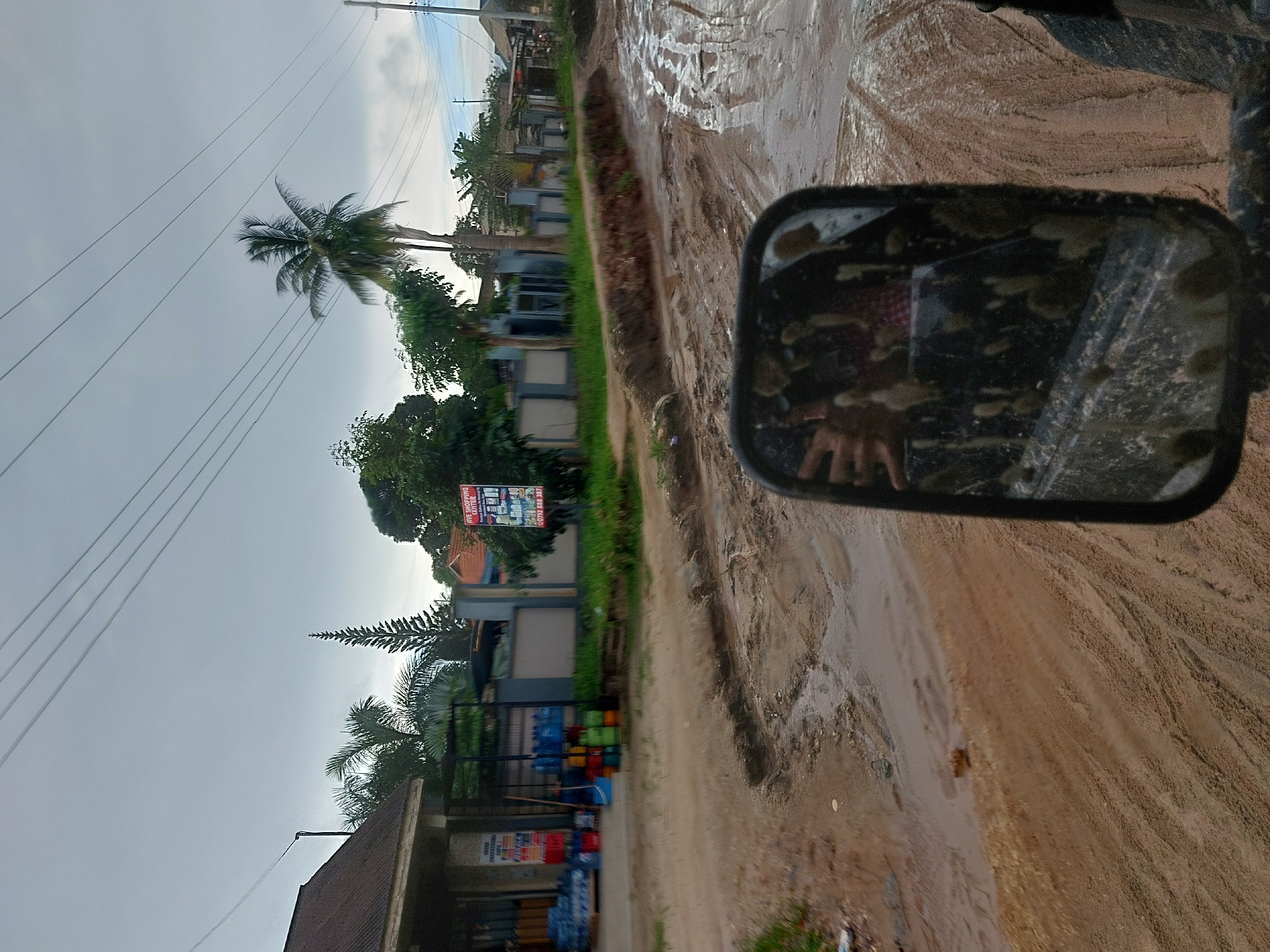 The image shows a muddy, dirt road in what appears to be a residential or commercial area. The scene is likely in Dar es-Salaam, Tanzania, based on the presence of tropical vegetation, including a tall coconut palm tree, and the types of building structures that can be seen. Power lines are also visible in the sky. A vehicle's side mirror is in the foreground, obscuring part of the view. The mirror's surface is dirty and shows a reflection of someone wearing a checkered shirt. A store with stacks of water bottles, stacked colored tubs, and a sign is also visible. The road itself is waterlogged, with muddy tracks and puddles. The sky is overcast.