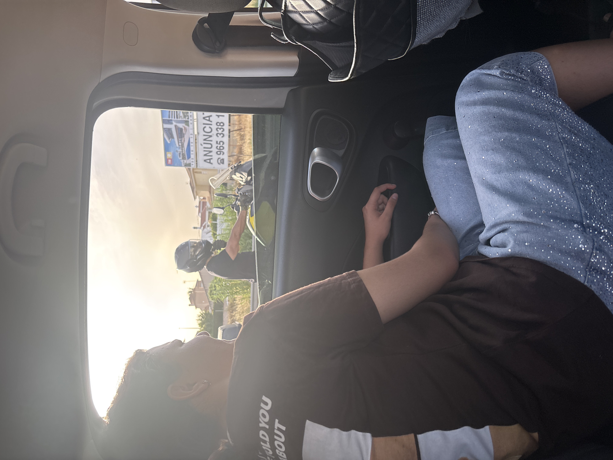 An image taken from inside a vehicle shows a person in the foreground wearing a brown t-shirt with white text that partially reads "OLD YOU ABOUT". They are also wearing light blue jeans embellished with small silver studs. To their left, another person is partially visible, wearing a white t-shirt. Outside the car window, a person wearing a motorcycle helmet is visible on a motorcycle. Behind them, a billboard displays an advertisement with Spanish text, including "ANUNCIA" and a phone number. The background shows buildings and vegetation under a bright, hazy sky, suggesting a sunny day. The car interior features a door handle and a speaker. The overall scene appears to be captured during a journey on a road.