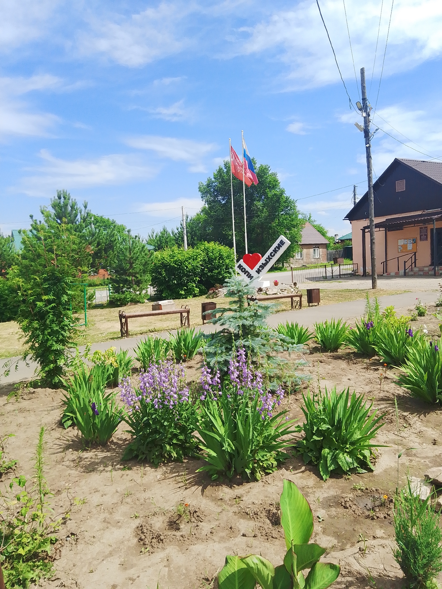 The image depicts a sunlit scene in the city of Изыхские Копи, Russia, featuring a decorative signpost, flags, and landscaping. The signpost, shaped like a diagonal bar with a red heart at the beginning, reads "КОПИ ИЗЫХСКИЕ" in white lettering, indicating the location. Two flags, one red and one with the Russian tricolor, are planted behind the signpost.
The environment is a public outdoor space, possibly a park or a town square. In the foreground, there is a well-maintained flower bed with lush green plants and clusters of purple flowers, interspersed with a small evergreen tree. A pathway, paved with light-colored material, runs through the scene.
Behind the flower bed, a brown wooden bench with decorative metal supports is visible. Further in the background, there are more trees, including pine trees and dense green bushes. A light-colored building with a brown roof and windows is situated on the right side of the image, likely a public facility such as a post office or administrative building, with a notice board on its exterior. A utility pole with electrical wires stands near the building. Residential houses with green roofs can be seen in the distance. The sky is blue with scattered white clouds, suggesting fair weather. No people are visible in the image.