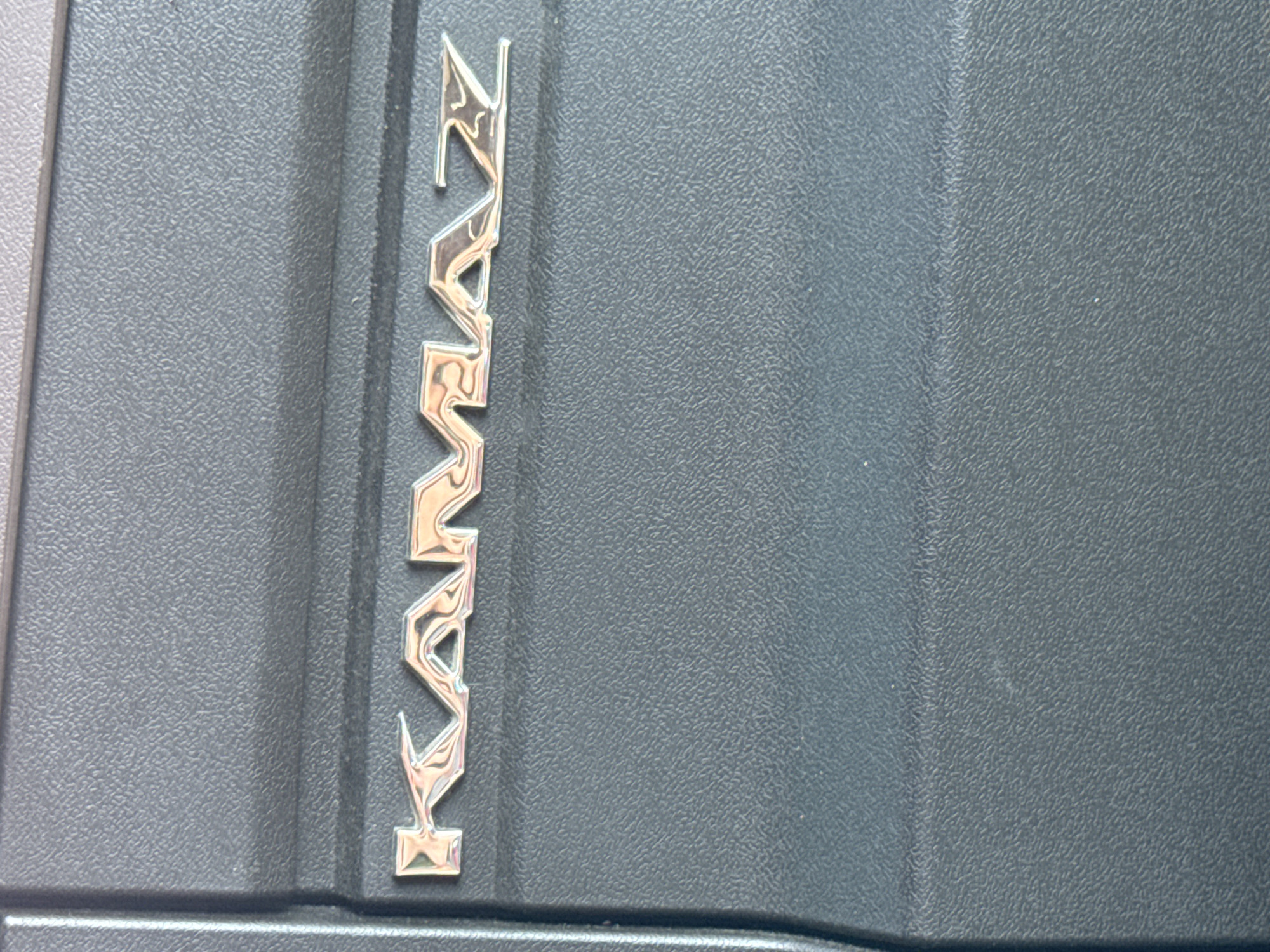 The image shows a close-up, vertical view of a silver metallic emblem with the word "KAMAZ" embossed on a textured, dark gray surface. The emblem is comprised of individual, stylized letters that appear to be chrome or polished metal, reflecting light and creating a shiny, three-dimensional effect. The "K" is at the top, followed by "A", "M", "A", "Z", and finally what appears to be a stylized "Z" or a symbol below it. The background surface has a fine, pebbled texture, characteristic of plastic or a similar material often used in vehicle interiors or exteriors. A subtle vertical seam runs down the right side of the frame, dividing the textured surface into slightly different planes. The lighting suggests an outdoor setting, likely daylight, given the bright reflections on the metal. The image is cropped tightly on the emblem and the immediate surrounding surface, providing no wider context of the environment or any people. The focus is solely on the branding of the vehicle. There is no discernible time of day, weather, or emotional cues. The location context is Sim, Russia, but this information is not visually evident from the image itself.