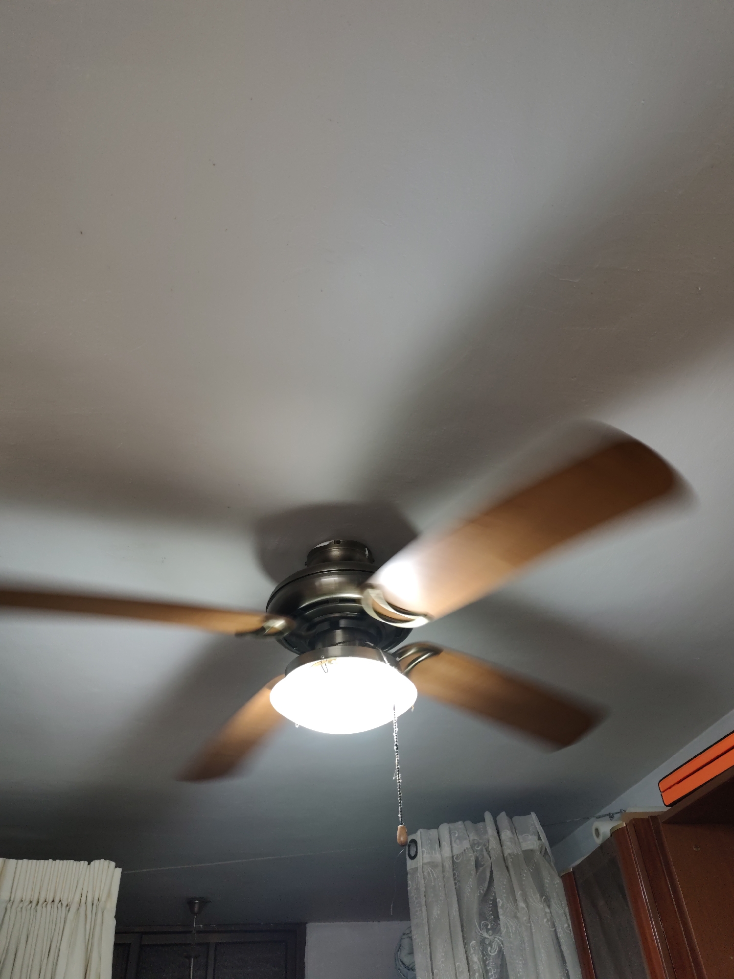 The image shows a close-up, upward-angle view of a ceiling fan with wooden blades, illuminated by its integrated light fixture. The fan is in motion, indicated by the blur of its blades. The ceiling is a plain, light gray color. Below the fan, a portion of a room is visible, including cream-colored curtains on the left, a translucent white curtain in the center, and a wooden cabinet on the right. The scene appears to be an interior residential space, likely a living room or bedroom, in the city of Or Akiva, Israel. There are no people visible in the image. The overall lighting suggests it might be daytime or a well-lit indoor space.