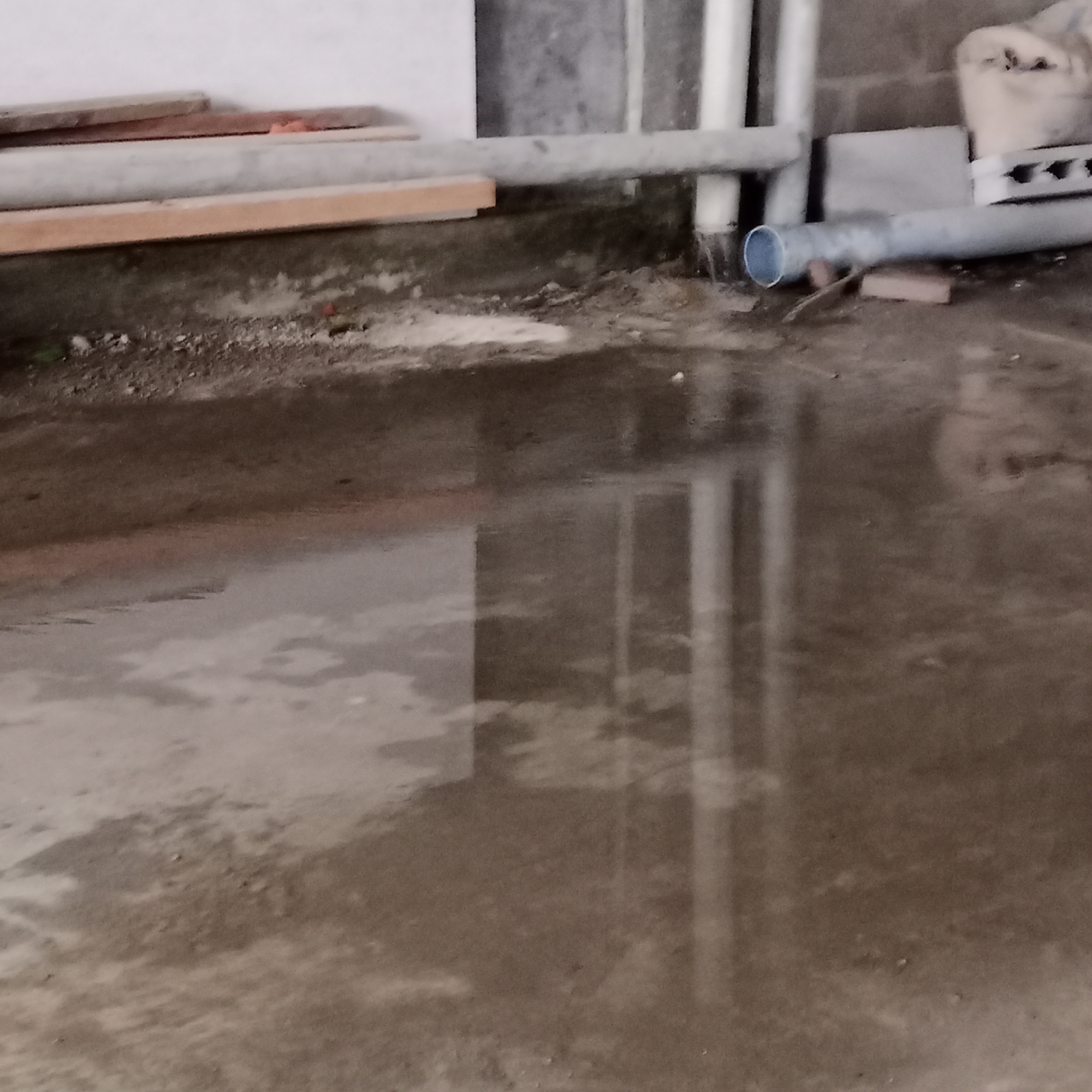 The image presents a ground-level view of a utilitarian or neglected area, likely outdoors or partially sheltered, in Lhokseumawe, Indonesia. The dominant feature is a large, murky puddle of water covering the foreground and a significant portion of the mid-ground. The water's surface reflects blurred images of vertical structures, primarily pipes and parts of a wall, suggesting a still body of water.
The ground around the puddle is a mix of dirt, sand patches, and concrete, with some visible debris and small pebbles. In the mid-ground, a horizontal gray pipe runs across the frame. Behind this pipe, against a textured, possibly concrete block wall, several vertical white pipes are visible. To the far right, another gray pipe, distinguished by a blue rim on its end, lies horizontally on the ground, appearing to be supported by a small brick. To the left of the frame, several wooden planks or boards are stacked against a lighter-colored wall.
The scene is devoid of people and shows no active event taking place. The presence of the large puddle strongly implies recent rain or a water leak. The overall lighting is diffused, making it difficult to ascertain the exact time of day, but there are no harsh shadows. The elements within the frame—pipes, construction materials, and the condition of the ground—suggest an industrial, utility, or construction-related setting. No visible text is present in the image.
