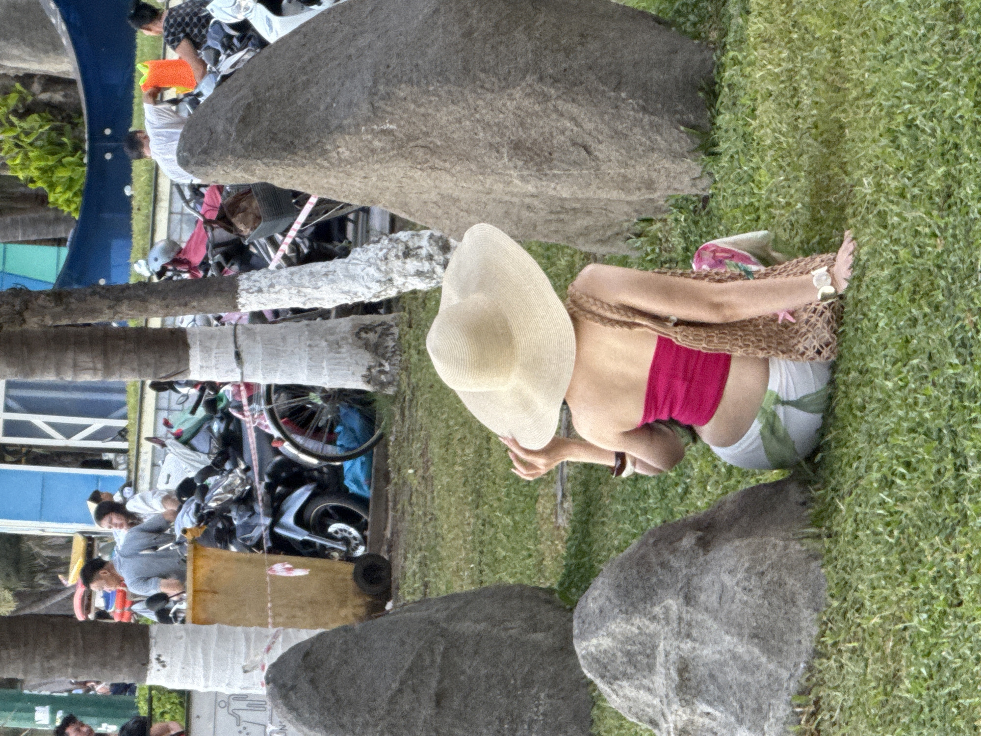 In Nha Trang Ward, Vietnam, a woman sits on a grassy area, facing away from the camera. She is wearing a wide-brimmed straw hat, a pink strapless top, and white shorts with a green leaf pattern. Her right hand is touching the brim of her hat. A woven bag rests beside her. Behind her, a variety of motorcycles are parked. In the background, several trees with white-painted trunks and lush green foliage are visible. Further back, a blue structure with a curved top and a white railing stands. People are seen in the background near the motorcycles and the blue structure. Large rocks are positioned around the grassy area where the woman is sitting.