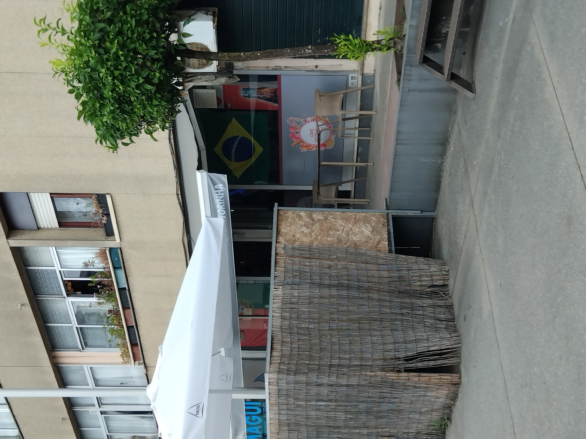 The image depicts the exterior of a commercial establishment, likely a cafe or restaurant, situated in Vila Nova de Famalicão, Portugal. The facade of the building features a large window displaying the Brazilian flag and a sign that reads "CAFÉ VITORINHA". In front of the establishment, a white umbrella provides shade over a patio area with tables and chairs. A bamboo-like fence partially obscures the entrance. The pavement in front of the cafe is made of concrete. The surrounding environment includes a tree with green foliage and part of a residential building. The scene is captured during the daytime, with natural light illuminating the area.