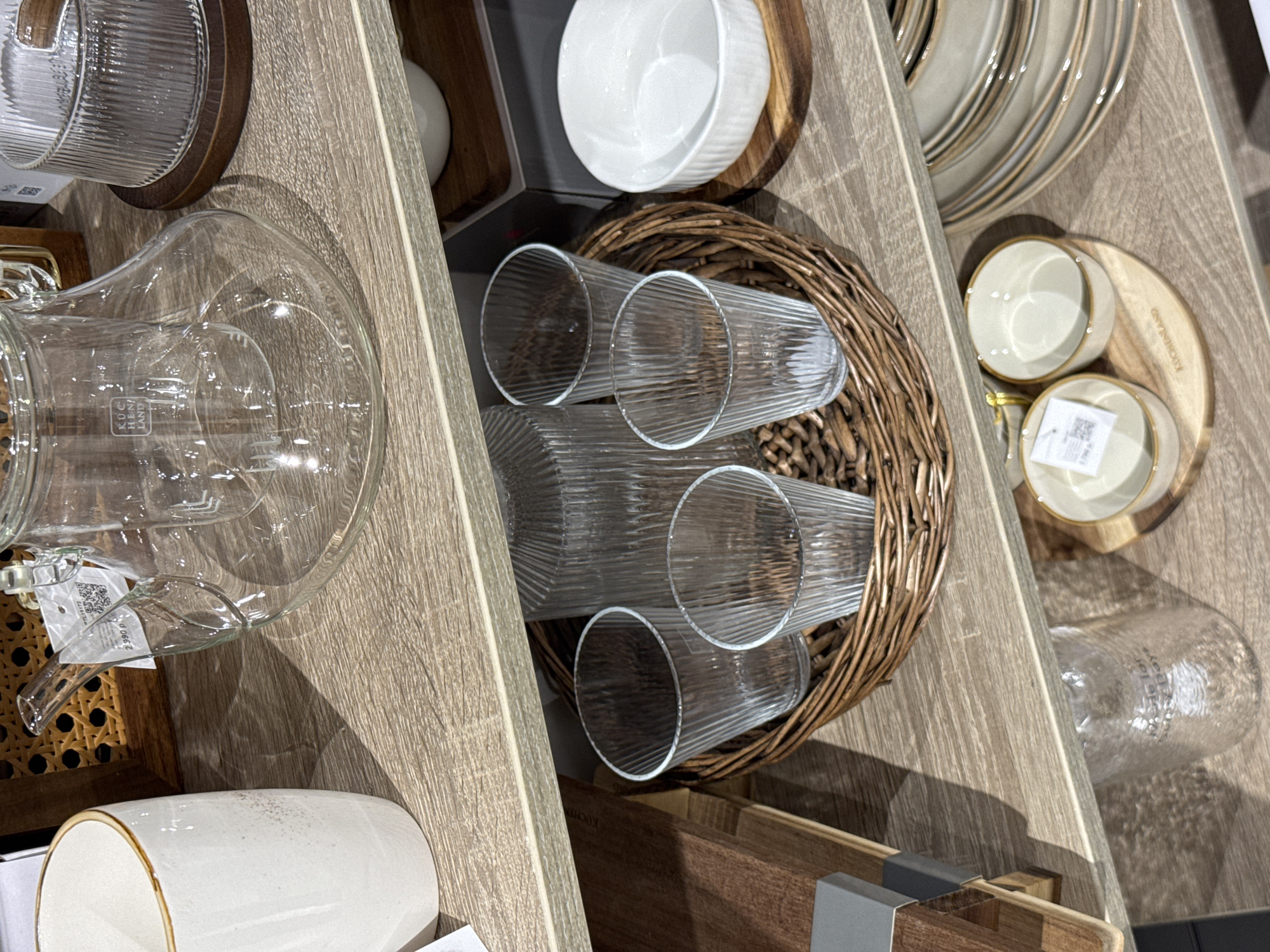 This image depicts three shelves displaying various kitchenware and tableware items in a retail environment, identified as a Kuchenland Home store in Perm, Russia, based on visible branding and currency. The shelves are constructed from light-colored wood laminate.
On the top shelf, a white ceramic mug with a light brown rim is positioned on the left. Centrally, a clear glass teapot or carafe, bearing the "KUCHENLAND" brand logo, features a clear handle and spout. A white price tag attached to the teapot handle displays "2 990 p" and a QR code. Behind the teapot, a section of a woven cane or rattan structure is visible. To the right, a portion of a vertically fluted clear glass item rests on a dark wooden coaster.
The middle shelf presents a round, woven wicker basket containing multiple clear glass items. These include a tall, vertically fluted glass bottle or carafe and four smaller, matching vertically fluted glass tumblers. To the right of the basket, a stack of white, vertically fluted ceramic bowls is situated on a round wooden cutting board or coaster.
On the bottom shelf, a clear glass jar with a textured, possibly rippled, surface is visible on the left, with faint text partially legible as "Made for Sunny Days". In the center, two small, light-colored ceramic bowls with light brown rims are displayed on a round wooden board, which is branded "KUCHENLAND". One of these bowls has a price tag showing "2 790 p". To the right, a stack of several light-colored ceramic plates or shallow bowls, also featuring light brown rims, is present. No individuals are visible in the scene.