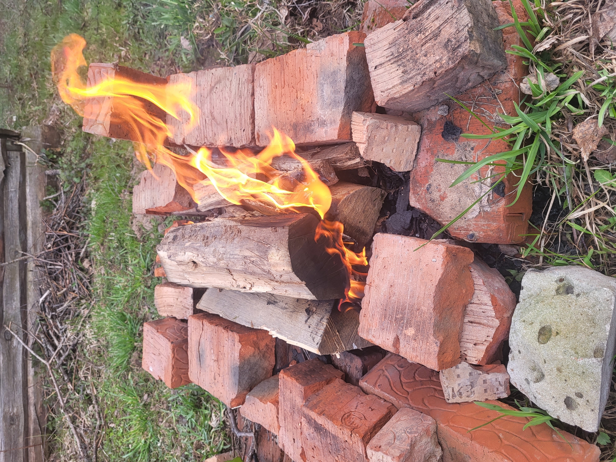 The image captures an outdoor fire burning on the ground, contained within a makeshift structure of bricks. Bright yellow and orange flames engulf several pieces of split firewood, casting a warm glow. The fire is surrounded by numerous reddish-brown bricks of varying sizes and conditions, some appearing worn or chipped, forming a loose perimeter. One larger, irregularly shaped grey stone with dark markings is also visible among the bricks on the lower right.
The setting appears to be outdoors, with patches of green grass, dry leaves, and bare twigs on the ground around the fire. A wooden fence or rough wooden structure is partially visible on the far left. The overall scene suggests a rural or semi-rural environment in Зипуново, Russia. The time of day appears to be daytime, possibly late afternoon, indicated by the natural light and the intensity of the fire's glow. No people are present in the image.
On a small, broken brick located towards the bottom right of the fire, the embossed characters "K3" are discernable. The fire appears to be a controlled burn, functional in nature, possibly for warmth or small-scale burning.