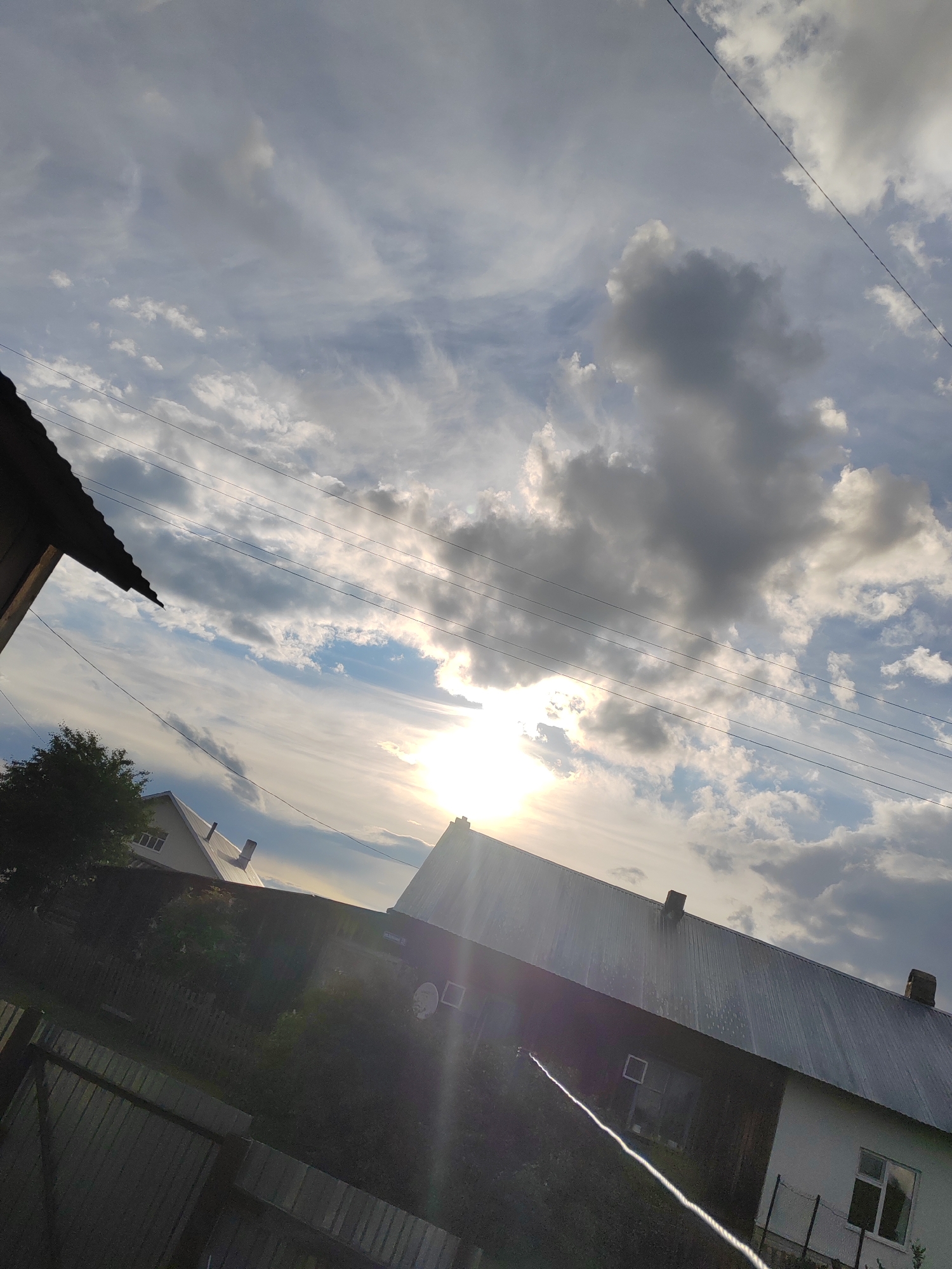 The image shows a daytime scene in Уральское, Russia. The sky is filled with a mix of white and dark gray clouds, partially obscuring the sun. Power lines stretch across the upper portion of the frame. Below the sky, a cluster of residential buildings is visible, including structures with metal roofs and chimneys. Vegetation, including trees and a wooden fence, is present in the lower portion of the image. A portion of a dark-colored structure is visible in the lower left corner. A metal fence is located at the bottom left. Sunlight creates lens flare effects.