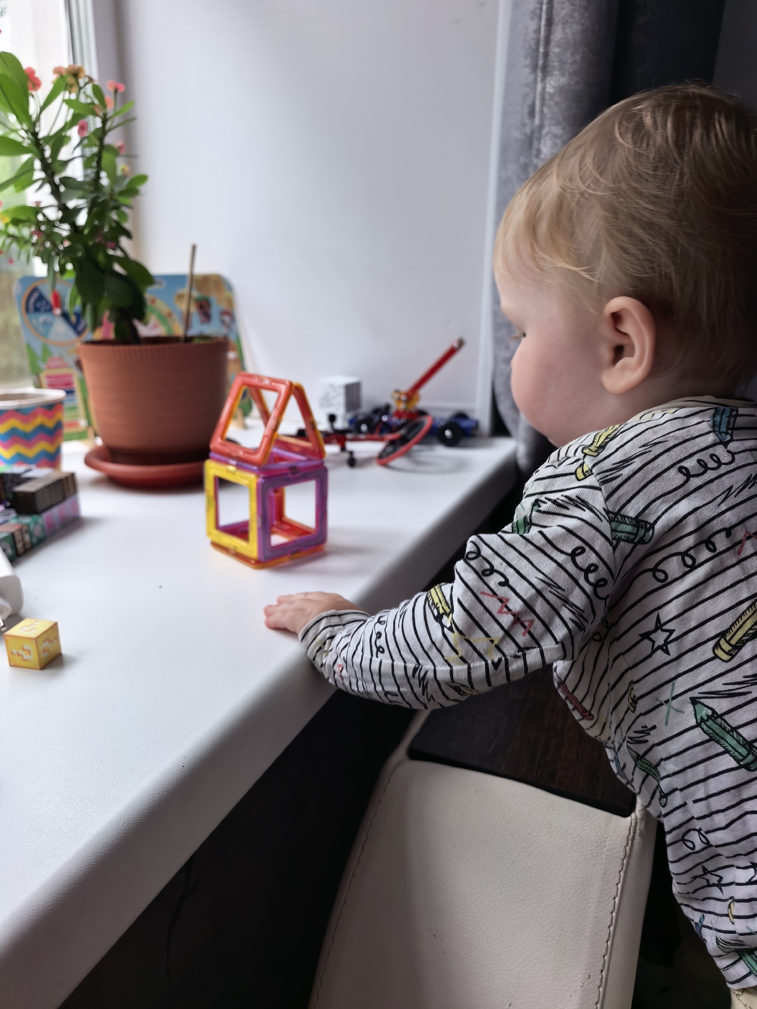 A blond-haired toddler, facing right, stands on a white surface, likely a windowsill or table, looking at magnetic building blocks. The toddler is wearing a black and white striped shirt with colorful drawings of pencils, stars, and scribbles. In front of the toddler is a small, multi-colored magnetic building block structure resembling a house, with yellow, purple, and orange pieces. To the left of the blocks, there is a plant in a terracotta pot, a colorful chevron-patterned cup, and other small toys and objects. The background shows a window with a gray curtain and a glimpse of the outdoors. The scene appears to be an indoor setting. There is no indication of the location being Sterlitamak, Russia.