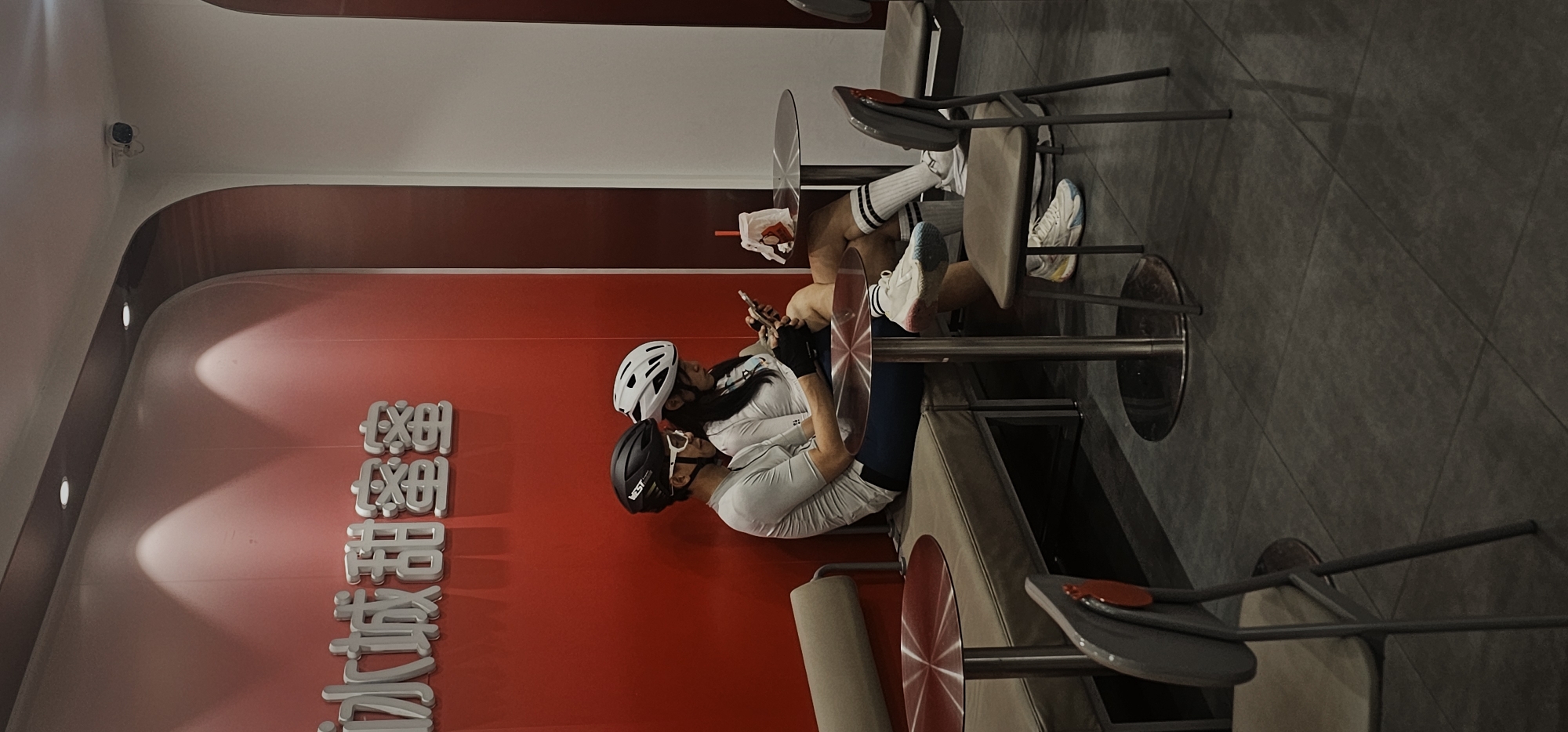 The image depicts an indoor scene in what appears to be a cafe or fast-food establishment, rotated 90 degrees clockwise. Two individuals, likely a man and a woman, are seated at separate round tables. They are dressed in cycling gear, including helmets (one black, one white), cycling jerseys, and shorts. Both are engrossed in their smartphones. One person has a beverage in a plastic cup with a straw on their table.
The setting features prominent red walls with a curved architectural detail near the white ceiling. Recessed lights illuminate this curve. The floor is tiled in a dark grey pattern. In the upper left corner, a security camera is mounted on the wall. The tables have metallic bases and circular tops, and there are chairs with light-colored upholstery.
Visible on the red wall are large, silver-colored Chinese characters. These characters spell out "蜜雪冰城" (Mìxuě Bīngchéng), which is the name of the popular Chinese tea and ice cream chain, Mixue Ice Cream & Tea. Above this, stylized characters resembling "MXBC" are also present, consistent with the brand's logo. The scene suggests a break during a cycling activity, taking place in a casual eatery in Jiulongpo District, China. The time of day is not ascertainable from the indoor lighting.
