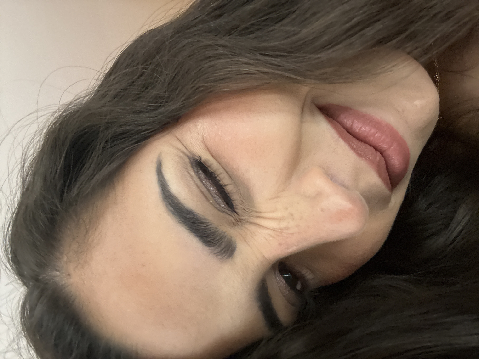 A close-up photograph features a woman's face tilted at an angle. She has dark hair, fair skin, and is wearing makeup, including eyeliner and lipstick. Her eyes are partially closed, and her expression suggests a slight grimace or wince. The background is out of focus and appears to be a plain wall. There is no indication of San Bernardino Tlaxcalancingo, Mexico, or any specific location information in the visible scene.
