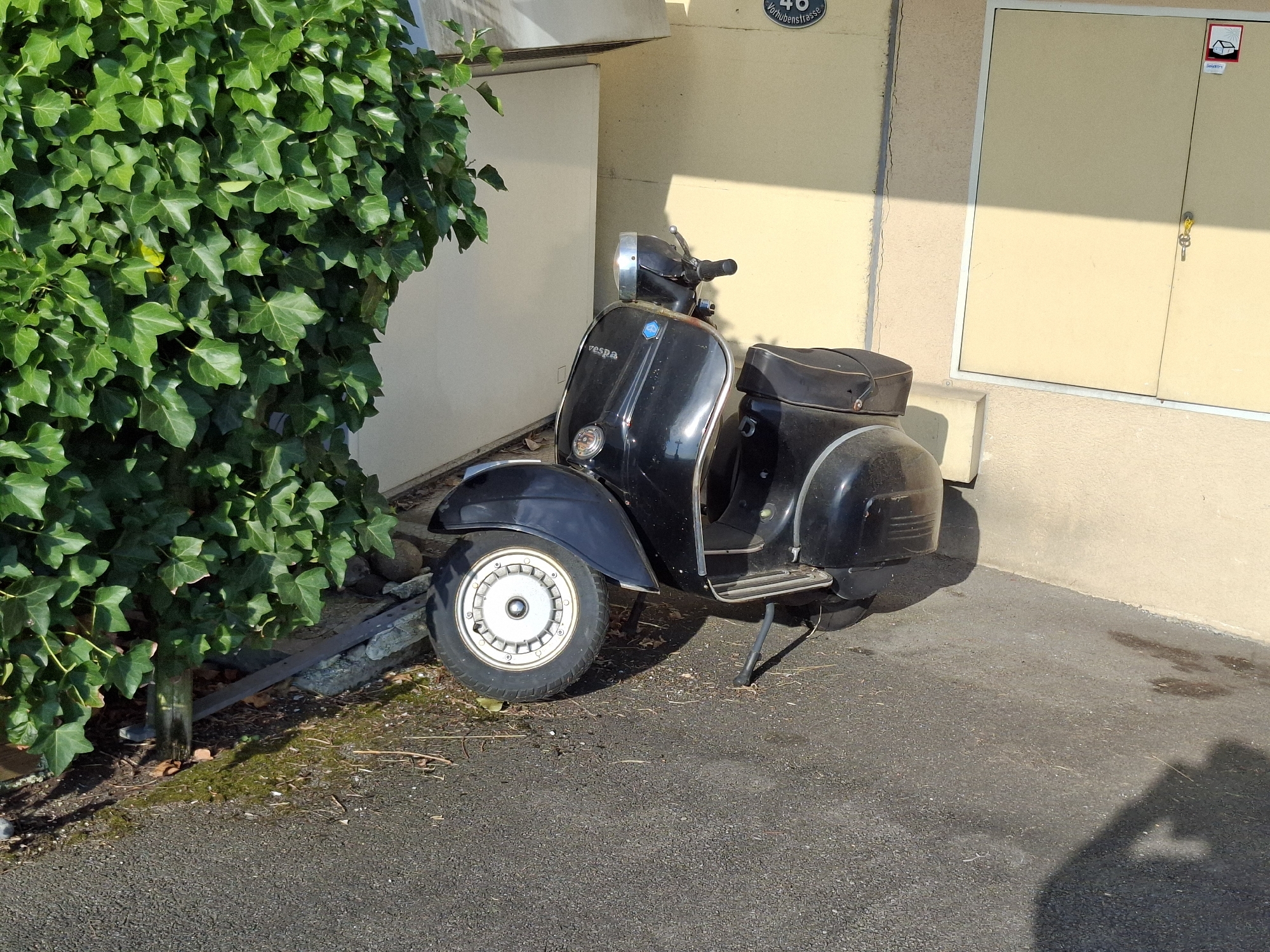 An outdoor, ground-level, medium shot captures a black Vespa scooter parked next to a building. The scooter is positioned facing right, with its front wheel angled slightly inwards. The building behind it is light tan in color and features a large, light yellow double door with a key in the lock on the right side. Above the door, a circular sign with the number "48" and text that reads "Vorhubenstrasse" is visible. To the left of the scooter, a dense, vibrant green ivy plant covers a significant portion of the frame. The ground is asphalt, with some scattered debris and patches of moss and dried leaves. Sunlight casts shadows from the scooter and the ivy onto the ground and the building. The scene appears to be a quiet, urban or suburban environment in Eschenbach (LU), Switzerland.