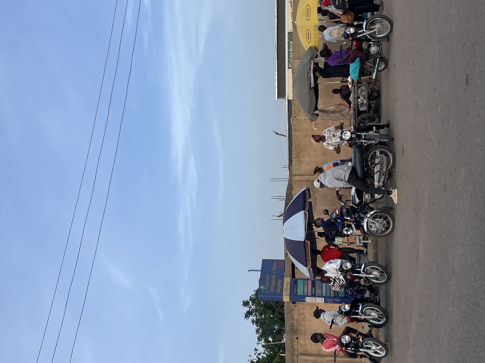 The image depicts a street scene, likely in Jos, Nigeria, based on the medical sign visible. Several motorcycles are parked along the edge of a road, with individuals either sitting on them or standing nearby. The road appears paved and is a light gray color.
A sign for "DaisyLand Orthopedic & Trauma Hospital" is visible, providing location context. The hospital sign is blue and displays hospital service offerings.
People are interacting around the motorcycles. Some are wearing casual clothing. Some people appear to be on waiting for a ride.
Additional commercial advertisements are visible, including a Heineken advertisement and MTN logos. The sky is clear with minimal cloud cover. Electrical wires run across the upper-left portion of the frame.
