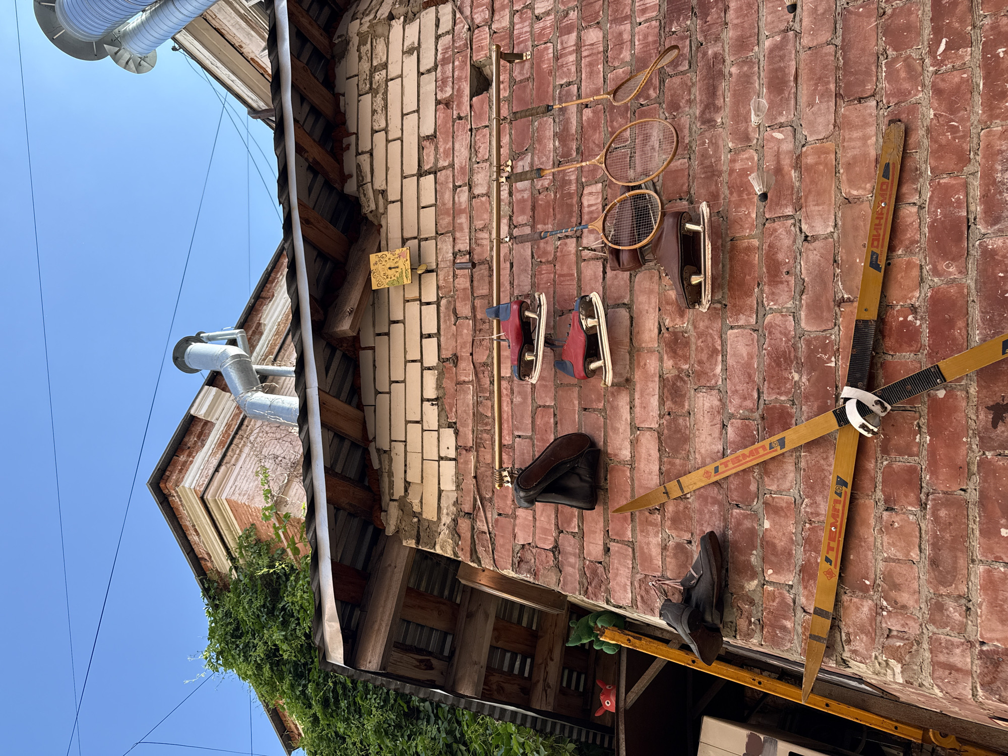 The image depicts an outdoor scene against a brick wall in Nizhny Novgorod, Russia. The wall is constructed of weathered red bricks, some of which are lighter in color. Several objects are mounted on the wall, suggesting a collection or display.
Prominently featured are a pair of wooden skis, positioned in an 'X' shape, with bindings visible. Above the skis, a rack holds various items. To the left, a pair of dark boots hangs by their laces. Next to them are three pairs of ice skates; two are red with white blades, and one pair is brown with metal blades. To the right of the ice skates are three wooden tennis rackets, two of which have intact strings, and the third has a visibly damaged stringing. Below the rackets and ice skates, several shuttlecocks hang from threads.
Further to the left, a small section of a building's roof is visible, covered with corrugated metal and supported by wooden beams. Green ivy climbs the side of the building, partially obscuring some of the upper wall. A yellow ladder leans against the wall to the left of the skis. Small, whimsical decorative items, including a red-faced ornament and a green plush toy, are visible on a shelf or ledge under the roof.
In the background, the blue sky is visible, along with overhead wires. The architecture suggests an urban setting, likely a courtyard or alleyway, typical of historical areas in Nizhny Novgorod. There are no people or animals present in the image. The overall impression is one of an eclectic outdoor display of sporting equipment and other items, possibly in a private backyard or an artistic installation.