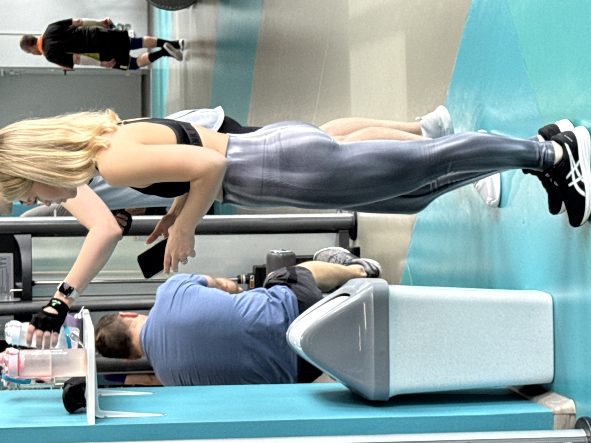 A photo shows a group of people at a gym. A woman in a black sports bra and gray leggings is in the foreground, bending over and holding a phone. She is wearing a watch on her left wrist and a black glove. A man wearing a blue shirt is near her, lying down and holding two water bottles. In the background, another man wearing a black shirt and shorts is doing an exercise. The gym has blue and gray flooring and equipment. This photo was not taken in Yekaterinburg, Russia, since the city's name is not displayed in any part of the scene.