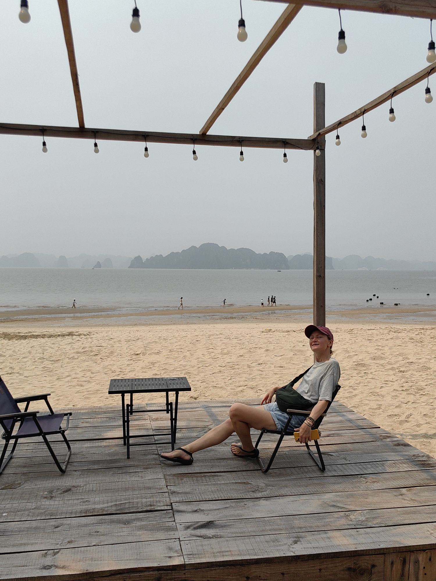 A medium shot reveals a woman reclining in a camp chair on a wooden deck at a beach. The deck features a second chair and a small, square table. The beach extends to the sea, where several people are visible. The sea has scattered islands in the distance. A wooden framework strung with lights hovers above the deck. The sky is overcast. The scene is in Bai Chay Ward, Vietnam. The woman wears a maroon baseball cap, grey T-shirt, denim shorts, and sandals. She also has a bag slung across her chest.
